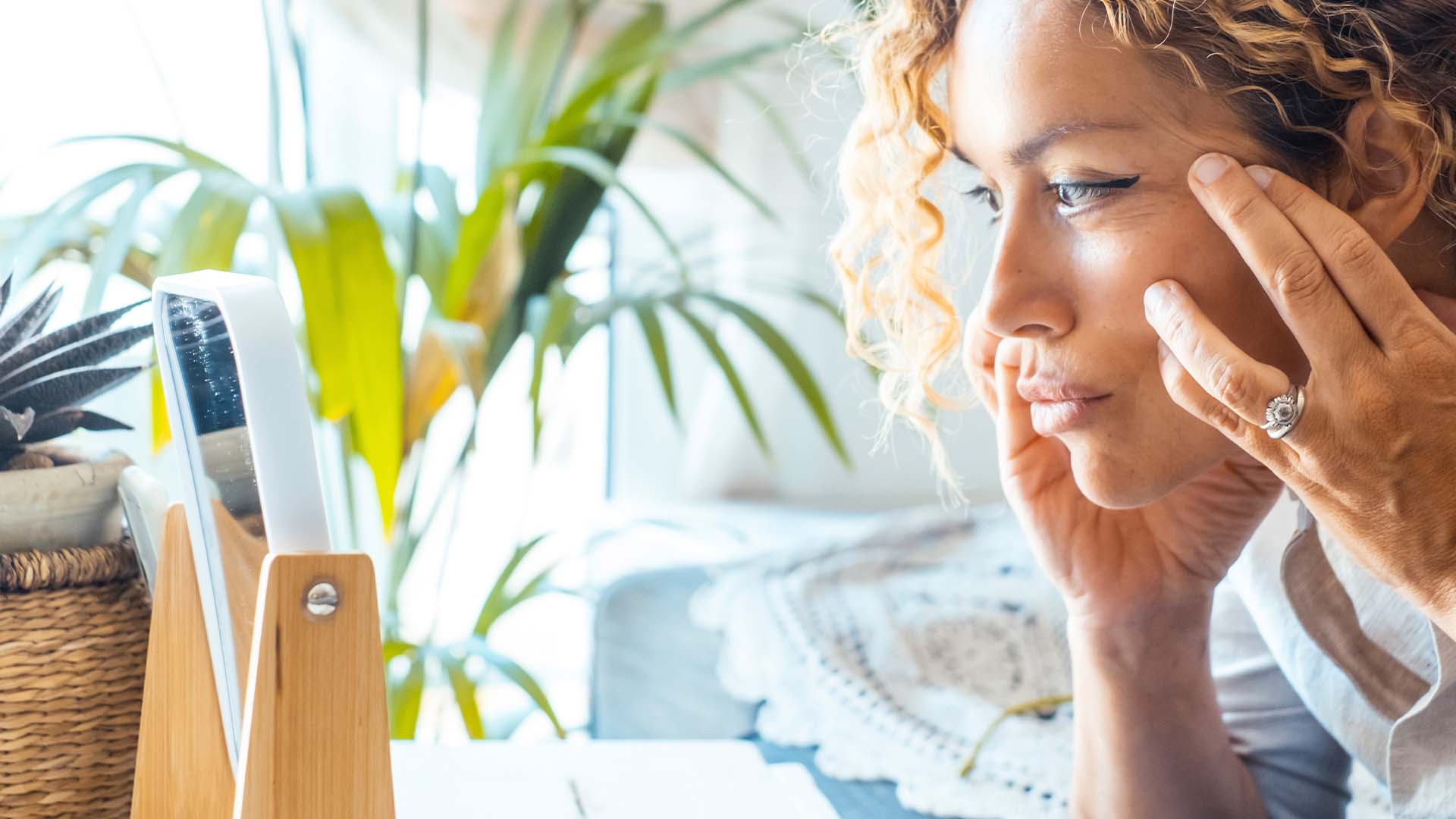 A woman thoughtfully looking at herself in a mirror while sitting in a bright room filled with greenery, symbolizing introspection and personal growth as part of managing depression with telehealth therapy.