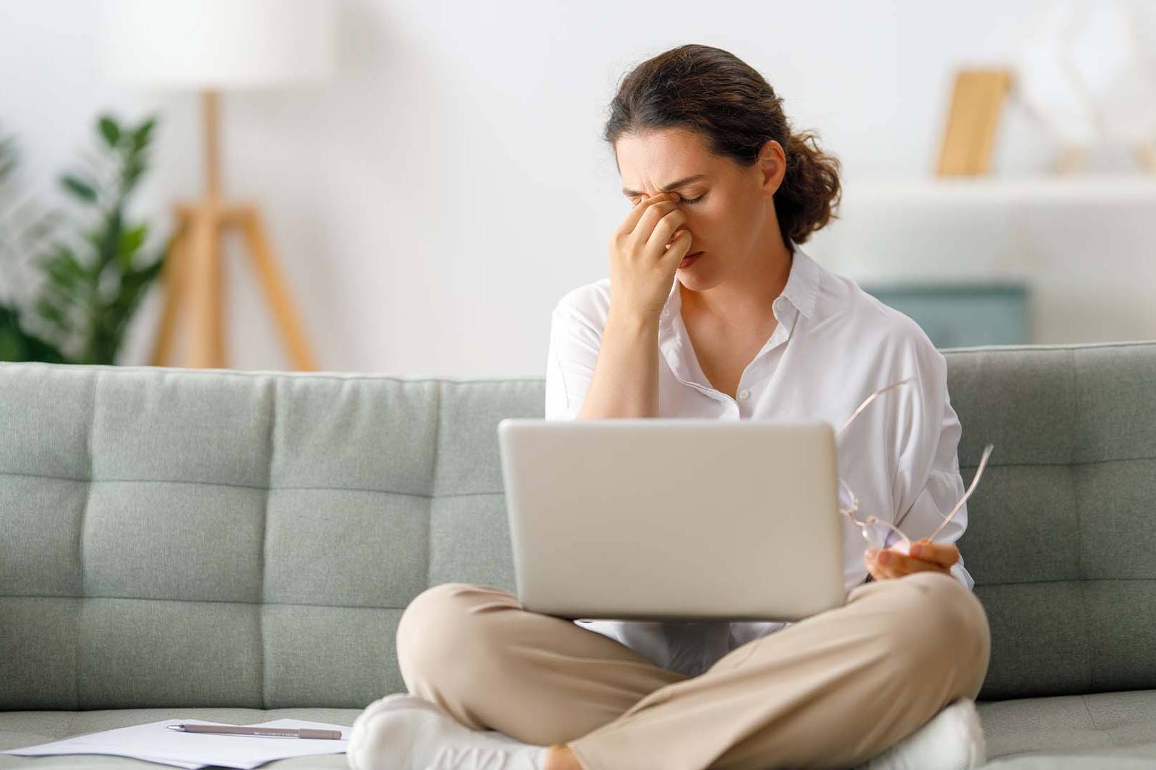 A woman sits cross-legged on a couch with a laptop on her lap, holding her glasses in one hand and pinching the bridge of her nose in a stressed gesture. The cozy home setting emphasizes the accessibility and comfort of online therapy for stress and anxiety.