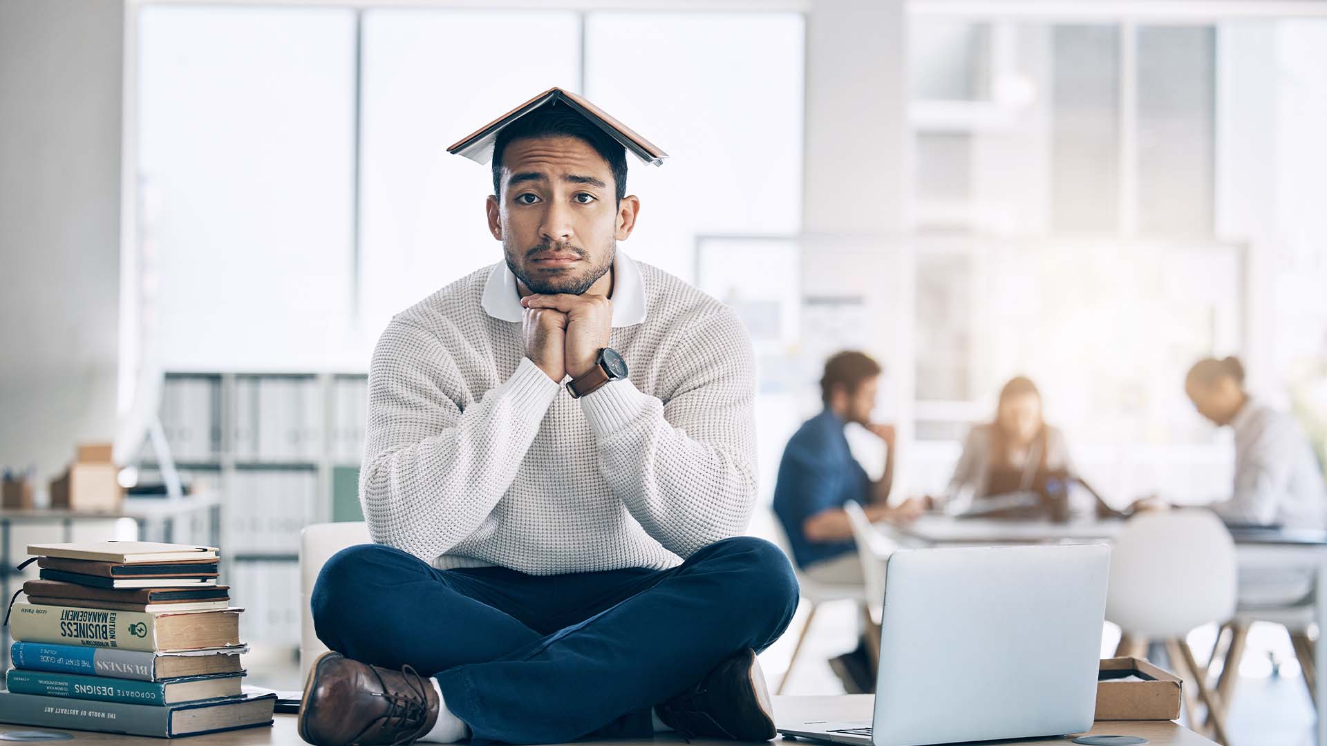 A young man sitting in an office with a book on his head, looking thoughtful and slightly overwhelmed, symbolizing the challenges of post-graduate life