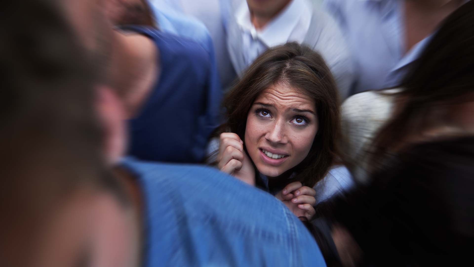 A woman appears distressed and overwhelmed in a crowded setting, clutching her hands close to her chest as she looks upward with anxiety. The blurred movement of surrounding people emphasizes her sense of isolation and discomfort.