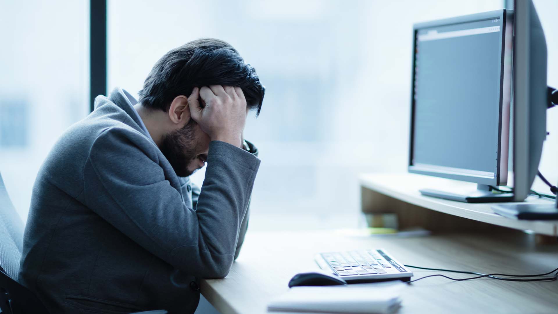 A stressed man with his head in his hands at a desk with dual monitors, representing workplace-related stress and depression
