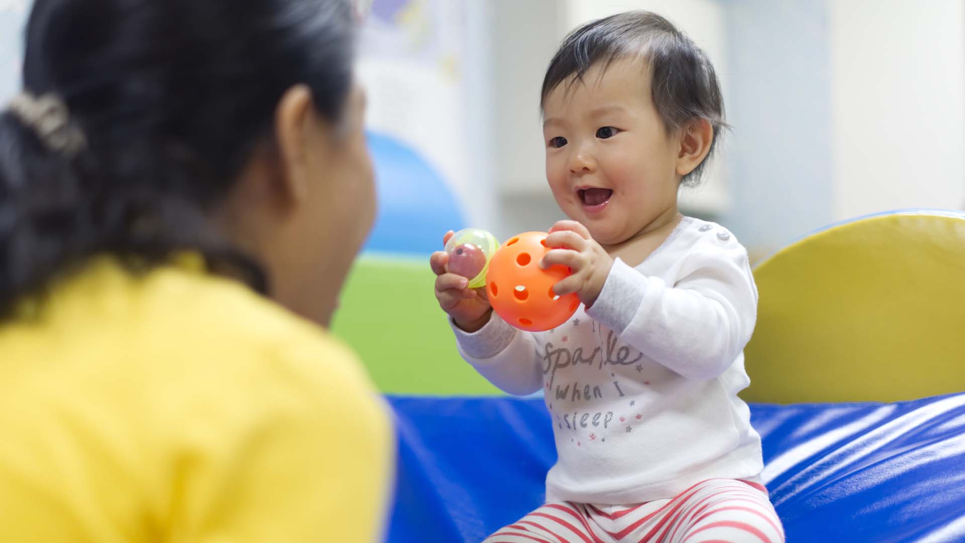 A happy baby playing with sensory balls during a developmental therapy session, guided by a therapist focusing on early cognitive and motor skills.
