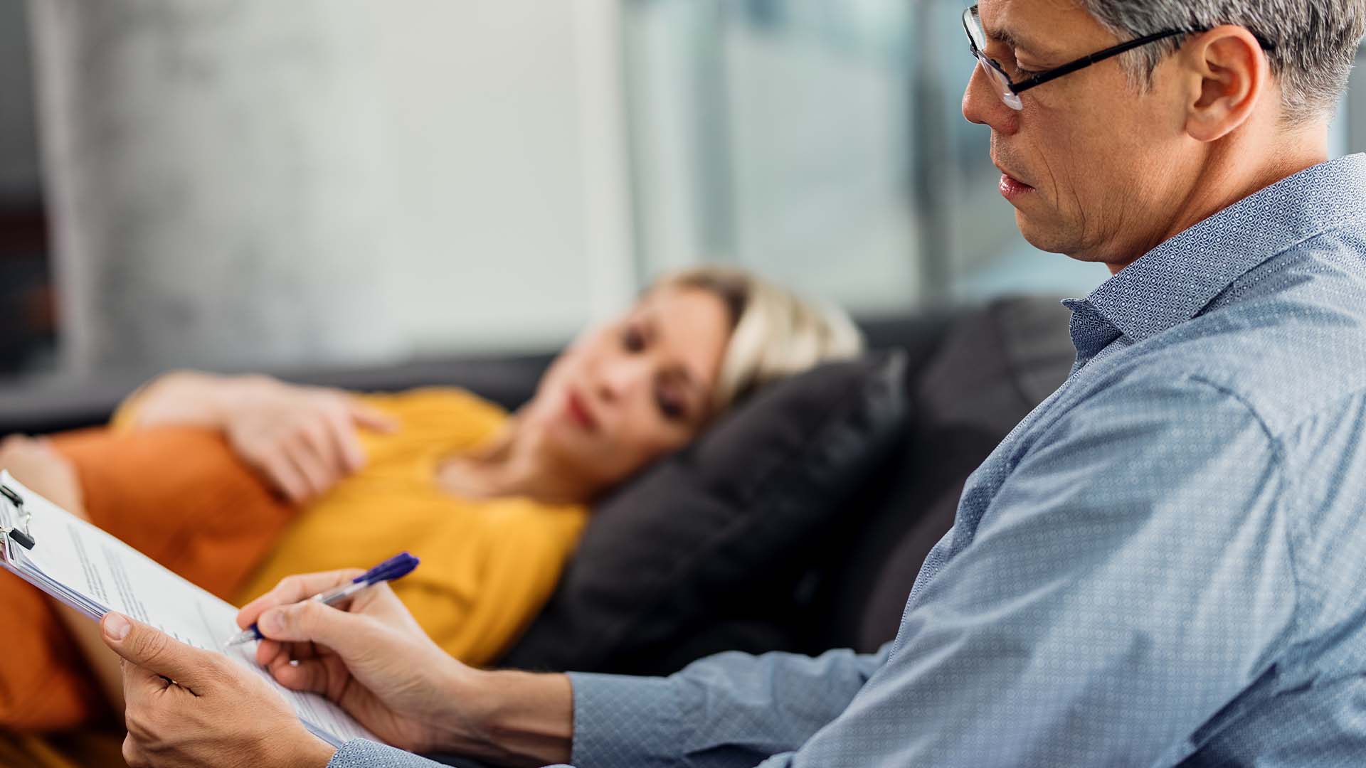 A therapist listens attentively while taking notes during a session with a client reclining on a couch, symbolizing the focus on relational interactions in FAP.