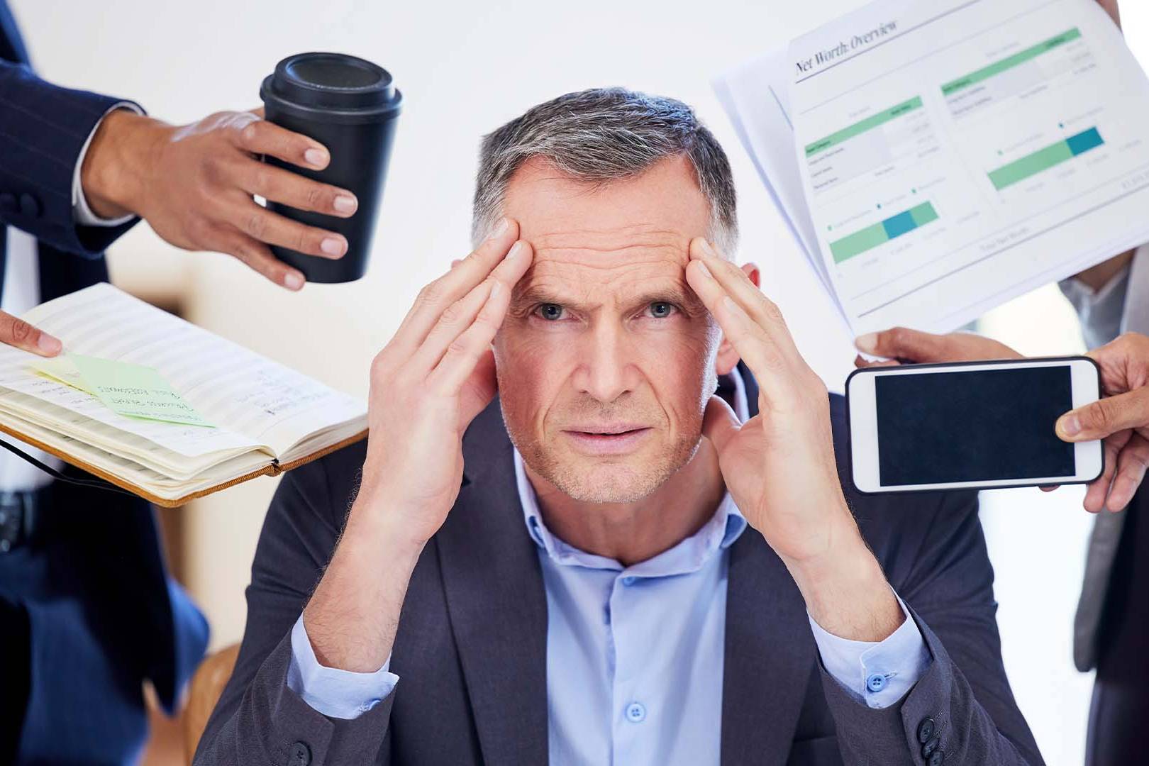 A stressed middle-aged businessman in a suit, holding his temples while multiple hands around him present a phone, coffee, documents, and a notebook