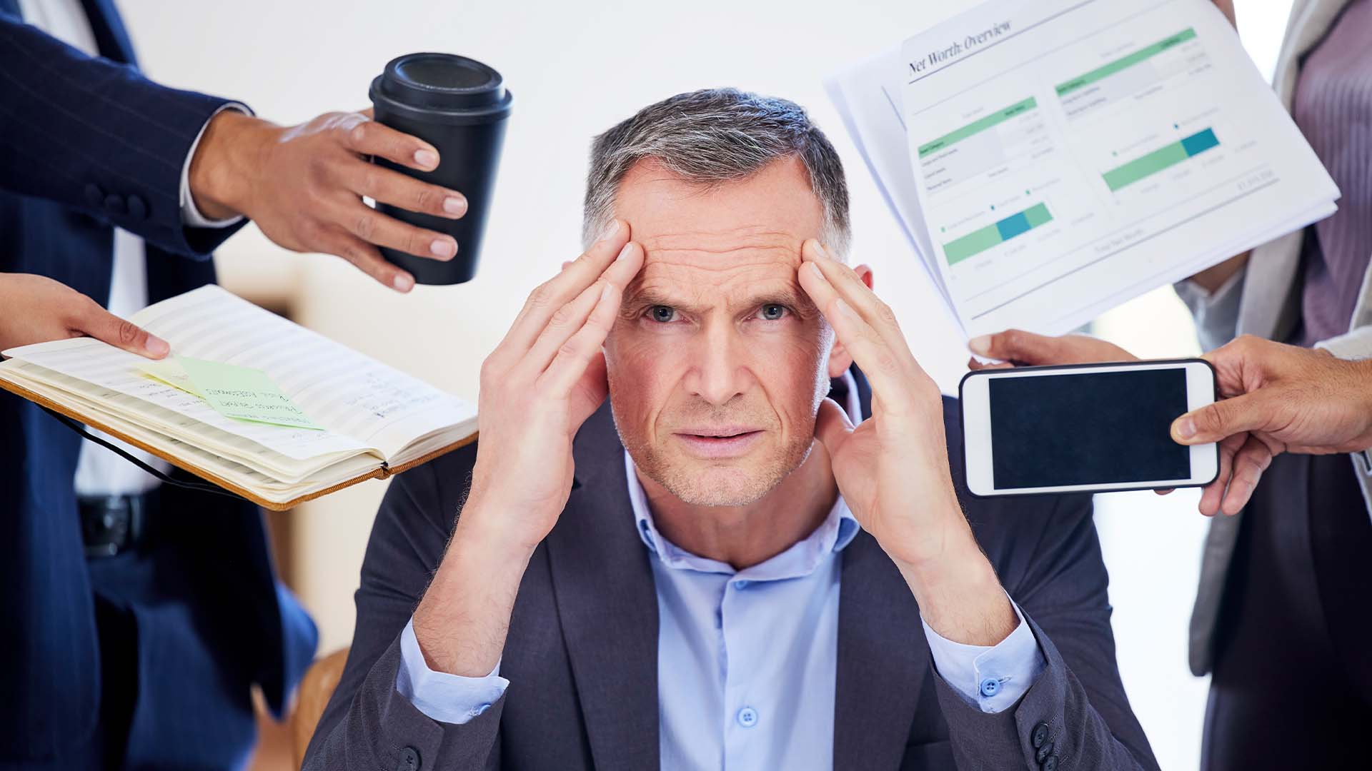 A stressed middle-aged businessman in a suit, holding his temples while multiple hands around him present a phone, coffee, documents, and a notebook