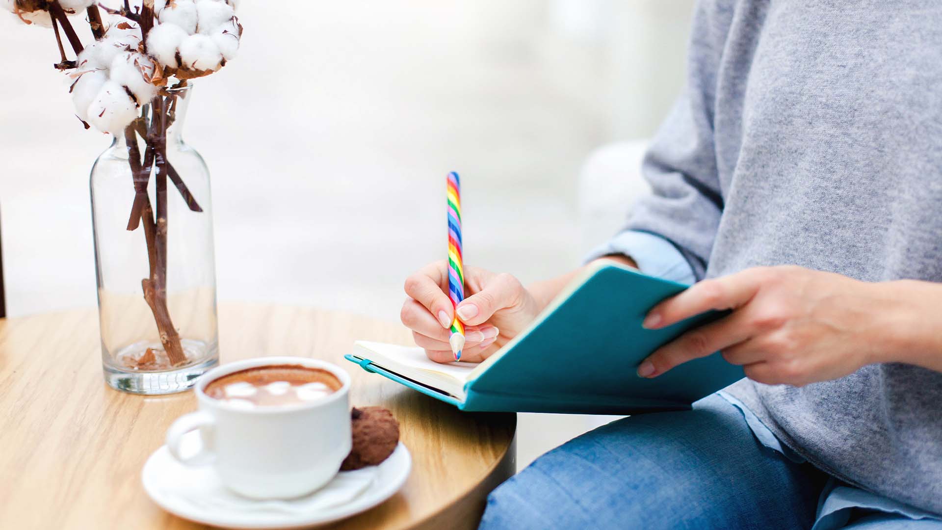 A person writing in a journal with a colorful pen, sitting next to a cup of hot cocoa and a vase of cotton flowers, symbolizing reflection and therapeutic writing.