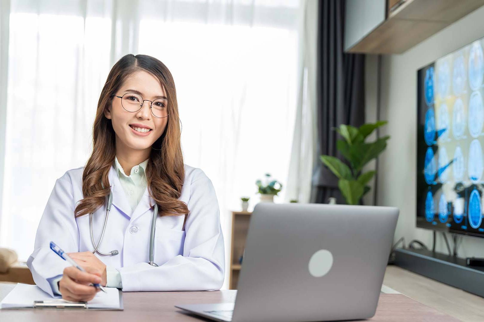 A smiling therapist in a professional setting during a telehealth session, with a laptop and brain scan images displayed on a monitor.