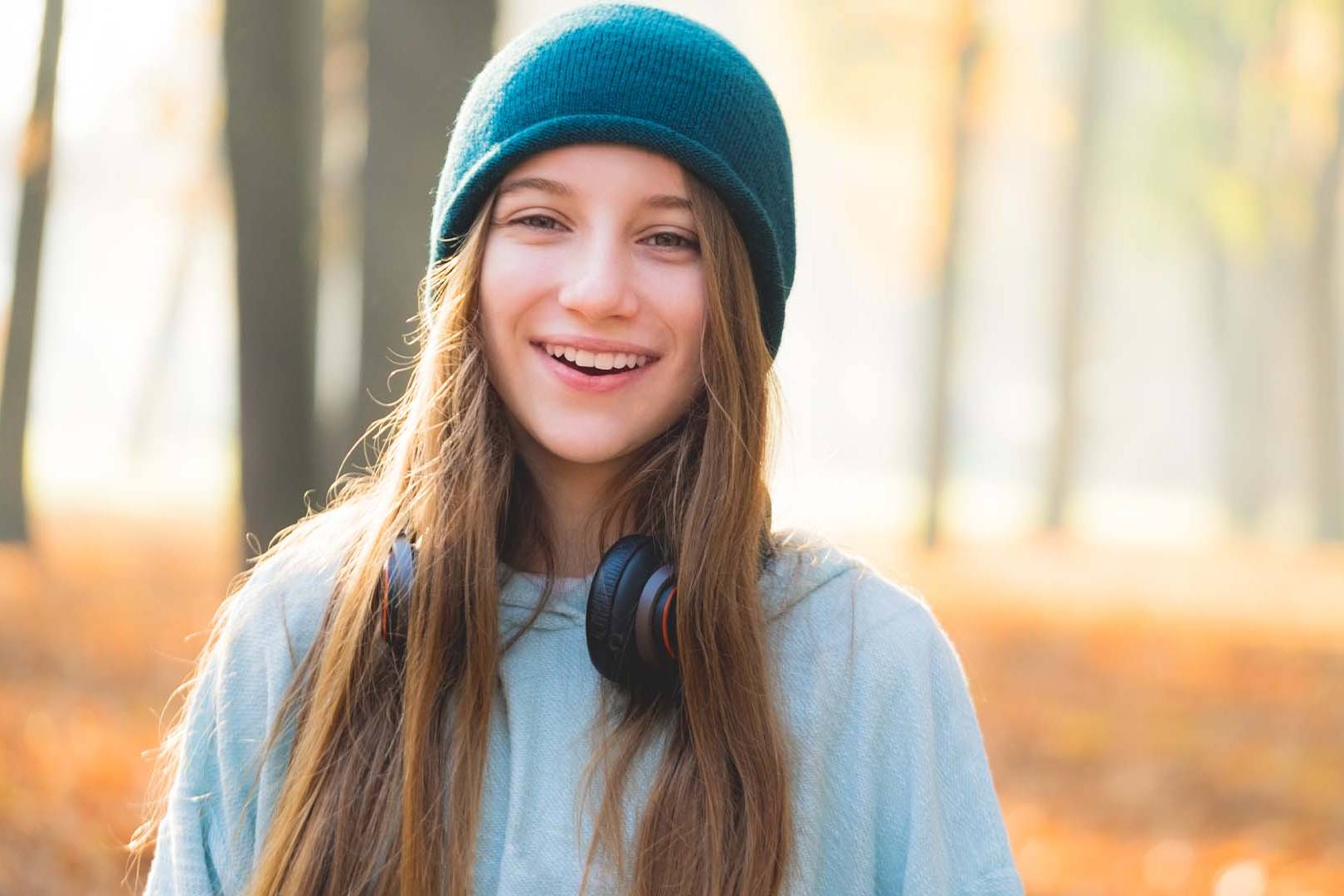 A smiling teenage girl outdoors in a forested area, wearing a beanie and headphones, radiating positivity and well-being.