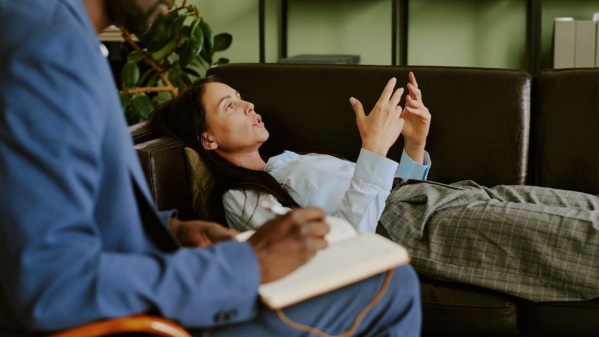 A woman lying on a black leather couch expresses her thoughts during a Metacognitive Therapy (MCT) session, while a therapist in a blue suit takes notes in a notebook.