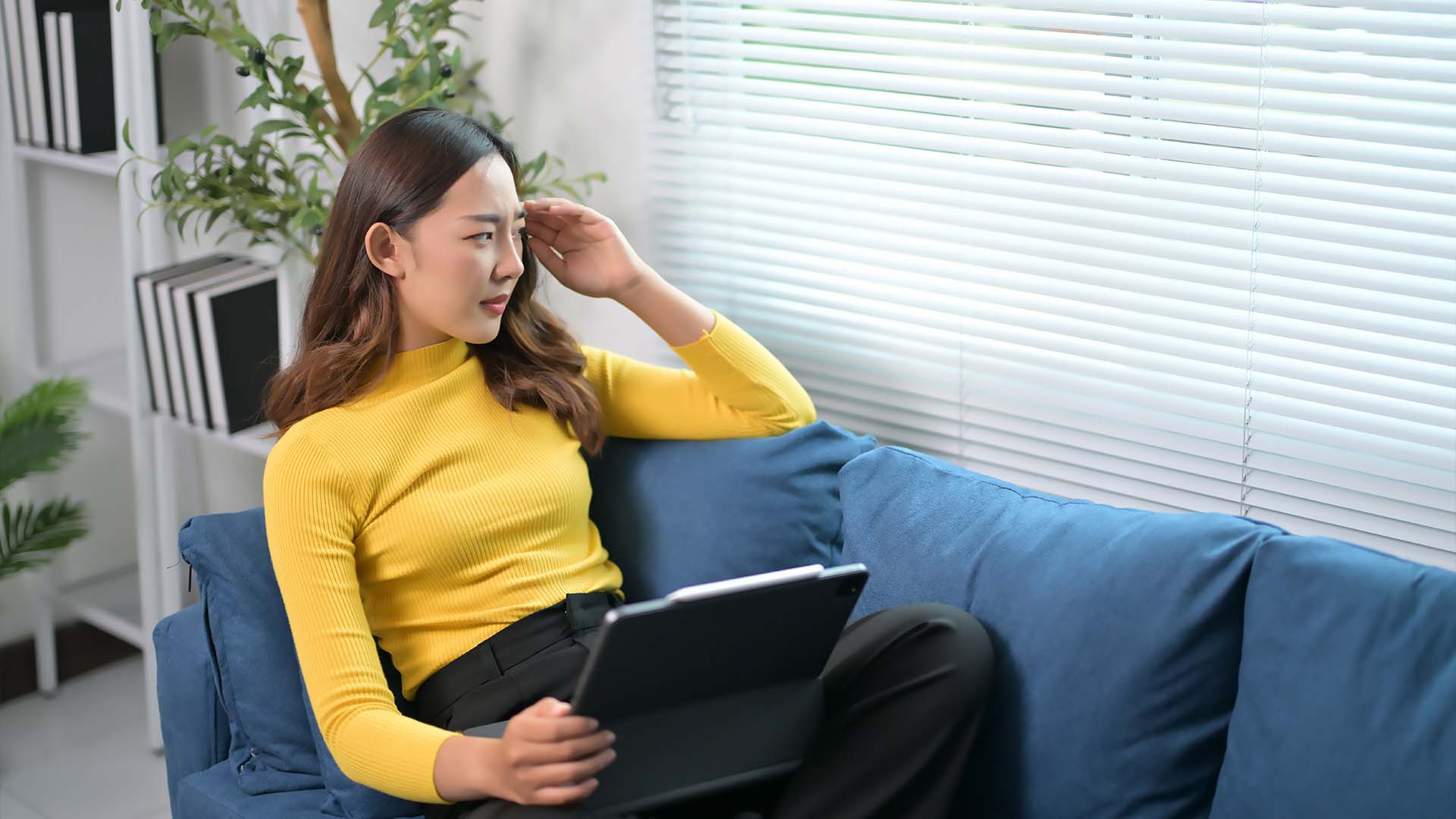 A woman in a yellow sweater sits on a blue couch, holding a tablet while looking concerned, with her hand resting on her forehead in a modern, well-lit office space.