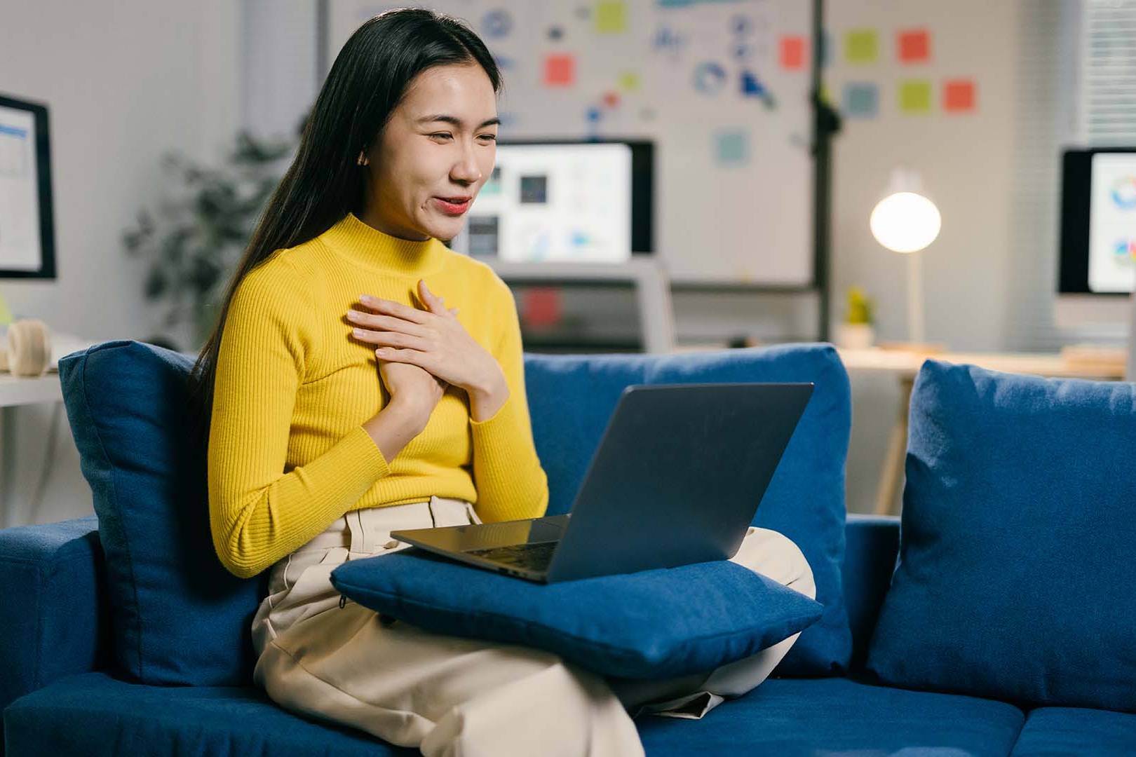 A woman in a yellow sweater sits on a blue couch with a laptop on her lap, placing her hands over her heart while engaging in an emotional virtual conversation in a well-lit office setting.