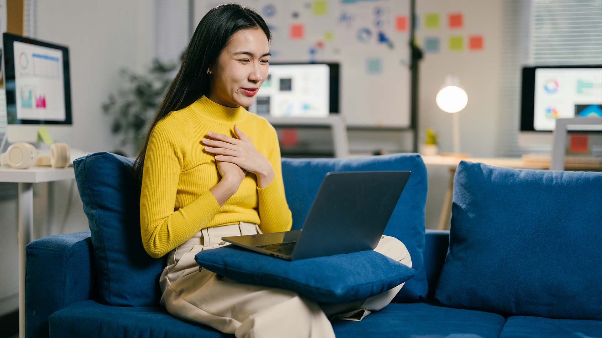 A woman in a yellow sweater sits on a blue couch with a laptop on her lap, placing her hands over her heart while engaging in an emotional virtual conversation in a well-lit office setting.