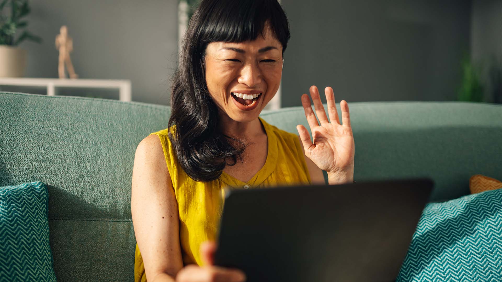 A woman in a yellow sleeveless top sits on a teal couch, smiling and waving during a virtual conversation on her tablet in a warmly lit living space.