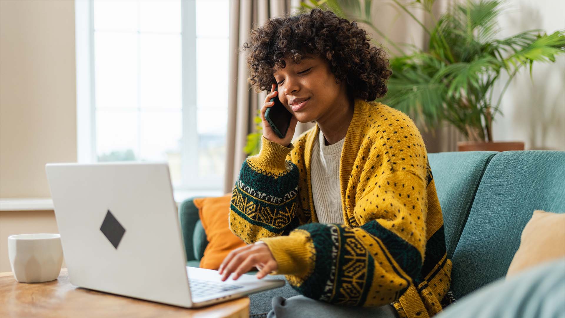 A woman in a cozy yellow and green cardigan sits on a teal couch, smiling while talking on the phone and using her laptop in a bright living room with plants and natural light.