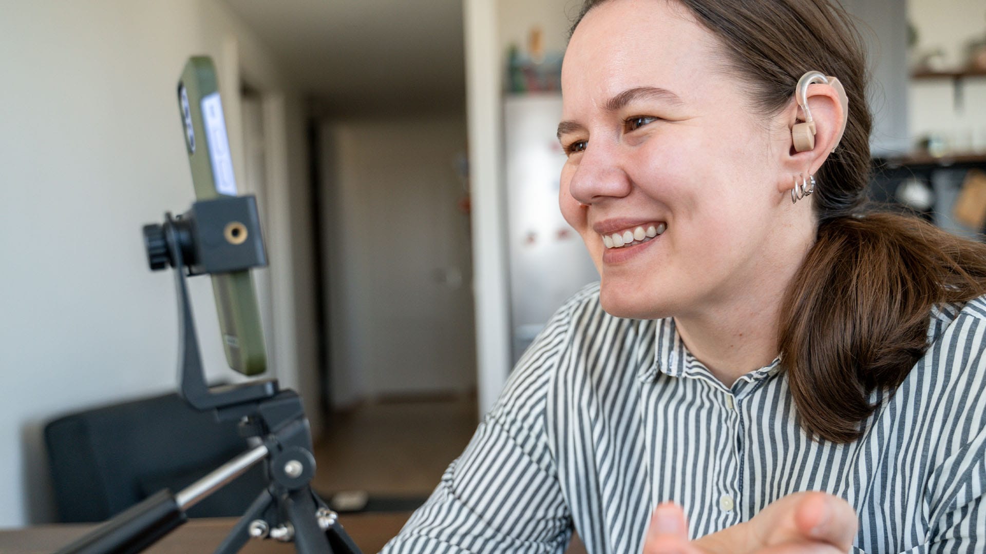 Smiling woman with a hearing aid participates in a video call using a smartphone mounted on a tripod, seated in a cozy, naturally lit home.