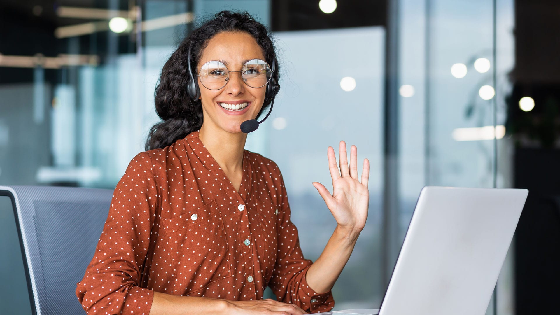 Smiling woman with a headset waves during a virtual session, seated at a desk with a laptop in a modern office space.