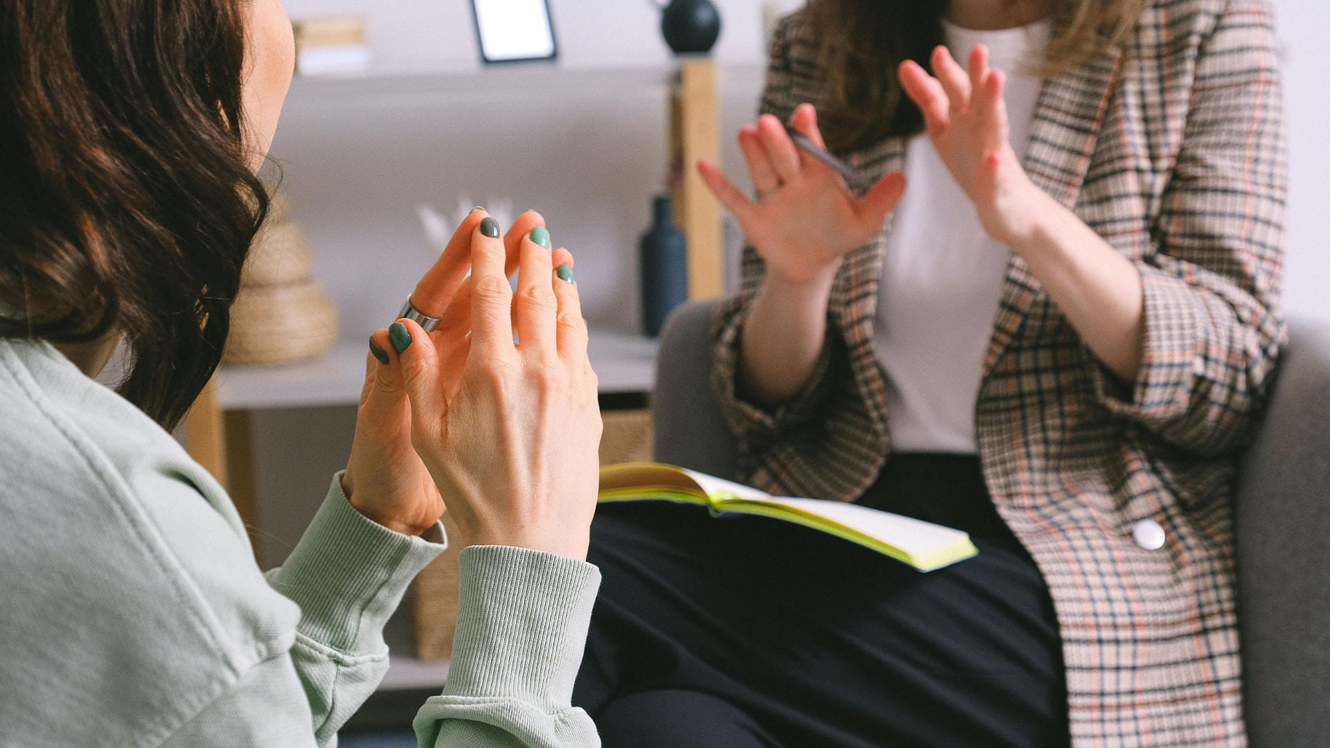 Two women in a counseling session use hand gestures while engaging in a thoughtful and expressive conversation. A notebook rests open on one of their laps.