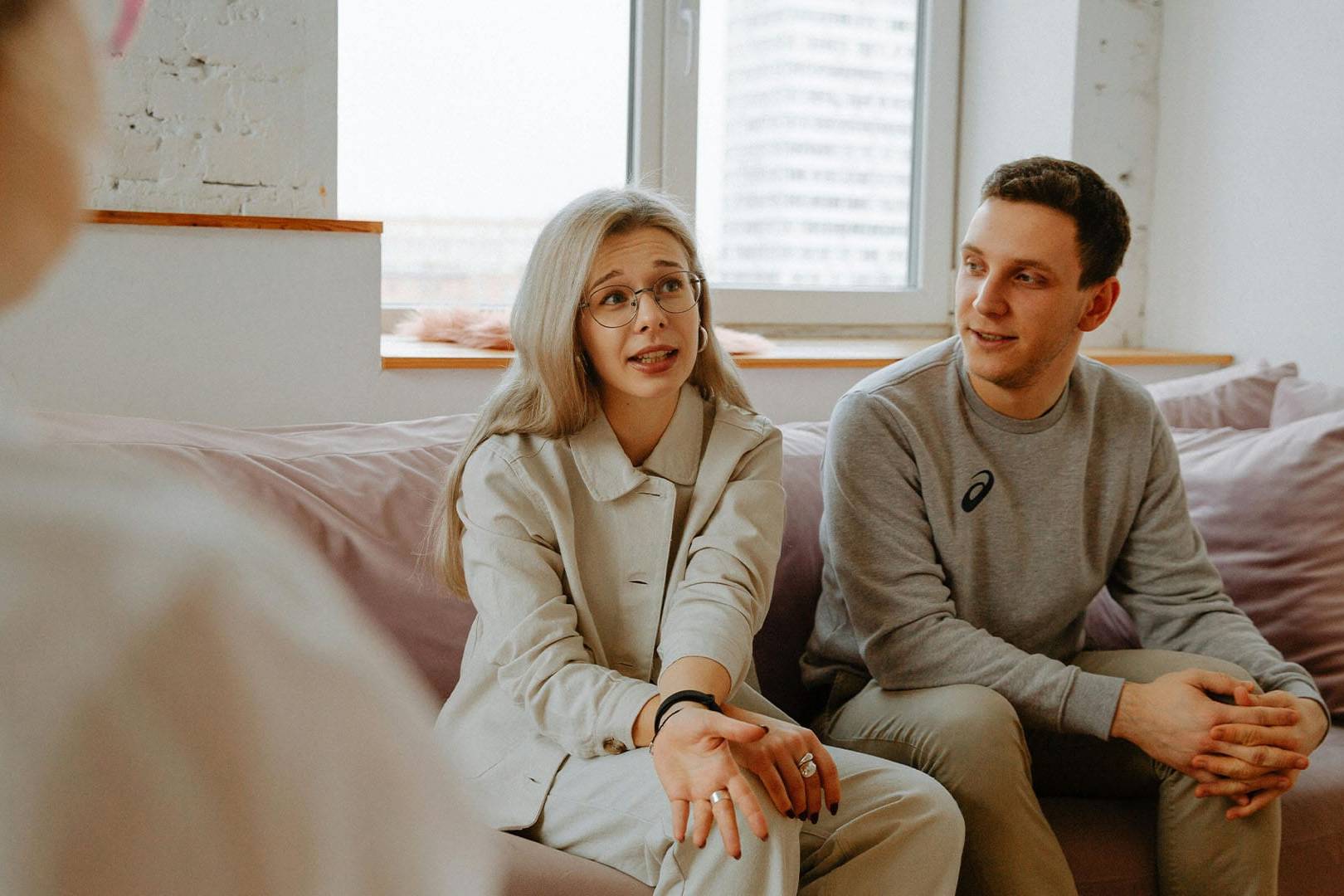 Couple engages in a therapy session, with the woman speaking and the man listening supportively on a couch in a bright room.
