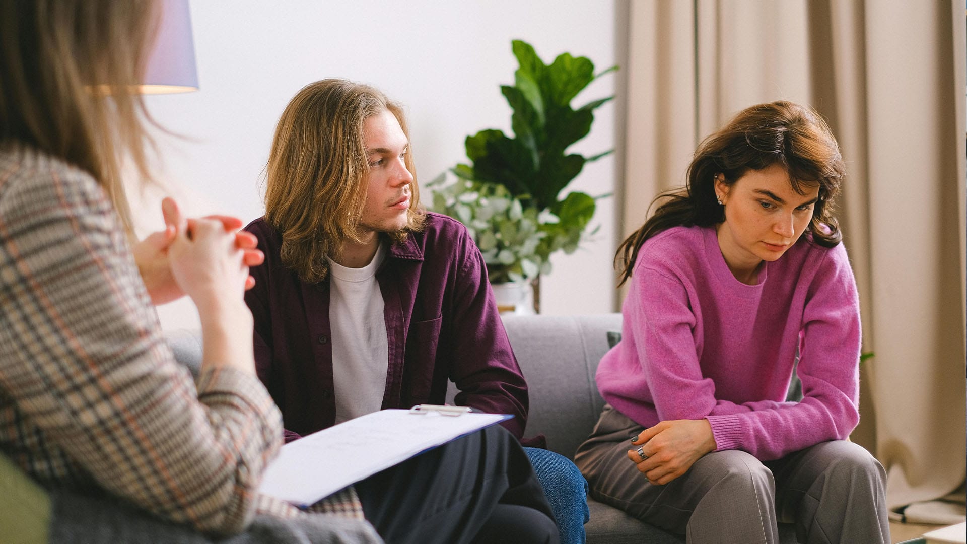 Couple in a therapy session with a clinician, both appearing emotionally tense while seated on a couch in a calm, professional setting.