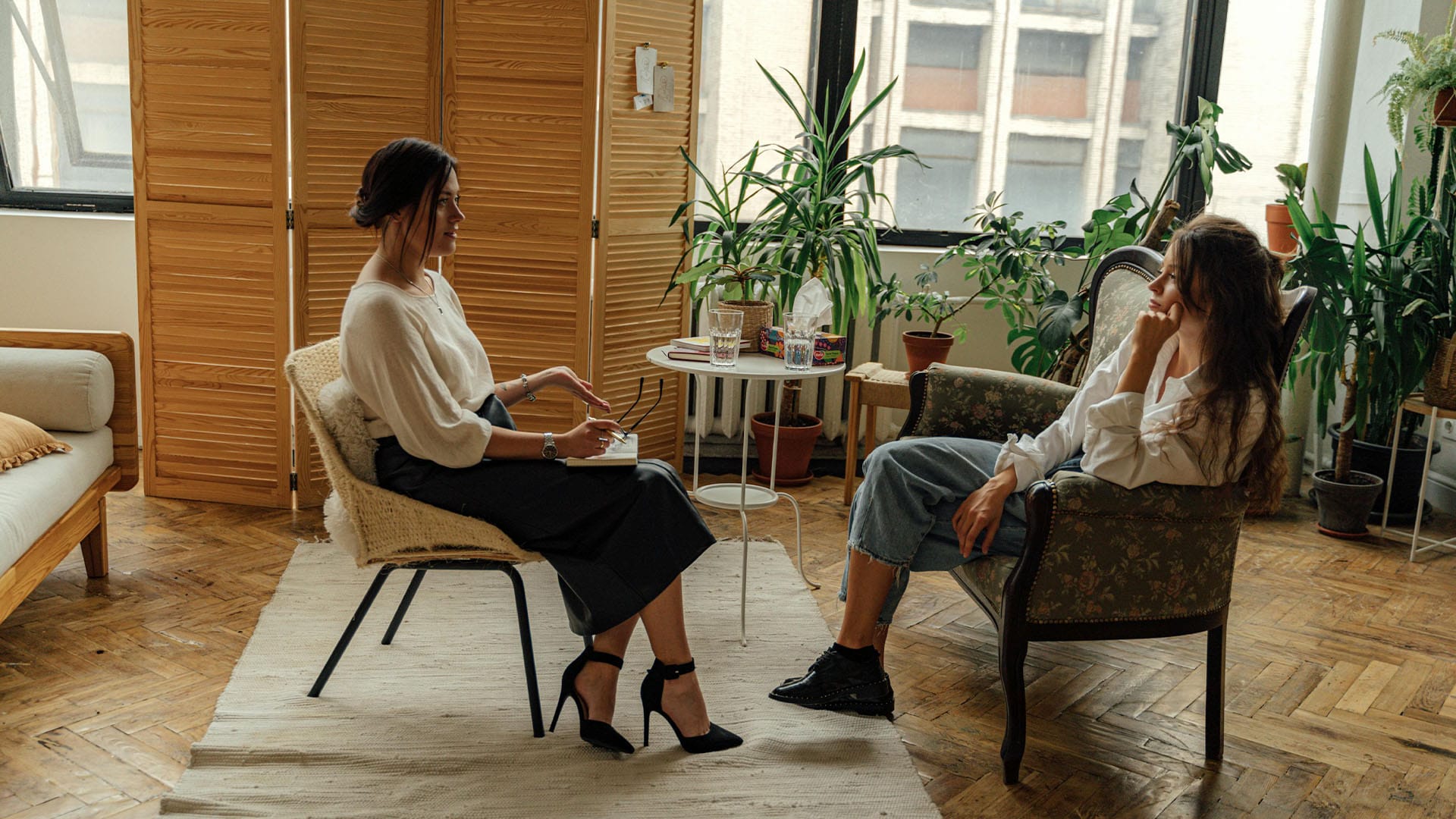 Two women in a warmly lit room with houseplants and natural textures engage in a therapy session—one appears to be the therapist taking notes, the other is seated reflectively.