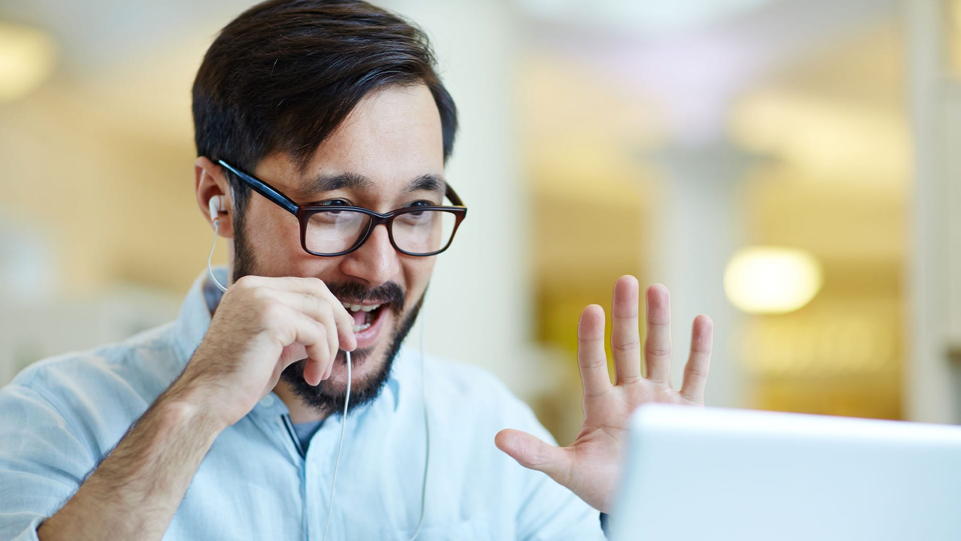 Man wearing glasses and earphones smiling and waving at his laptop during a virtual call.