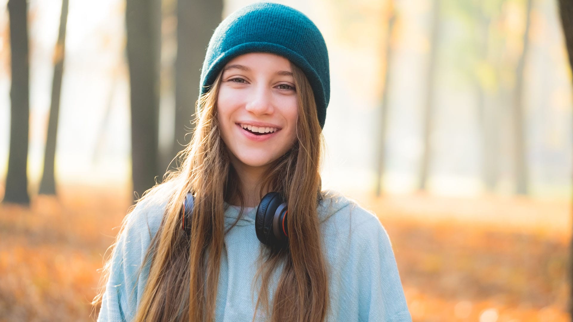 Smiling teen girl wearing a teal beanie and headphones around her neck, standing in a sunlit forest during fall.