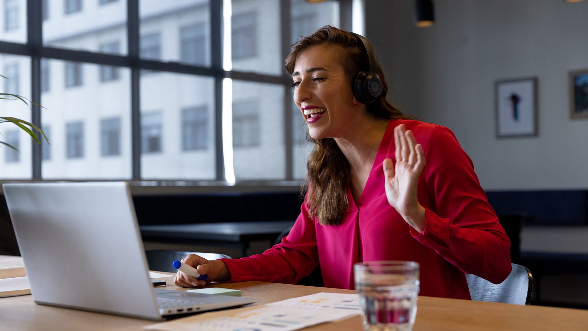 Woman smiling and waving during a virtual session, wearing headphones and sitting at a desk with a laptop.