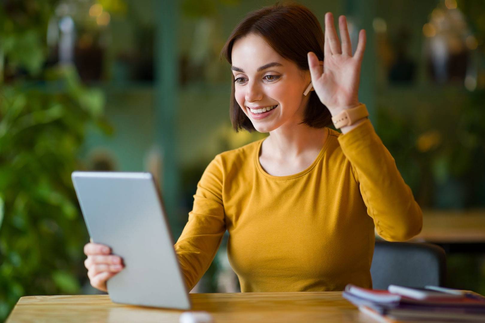 Smiling young woman with short hair waving at a tablet during a virtual meeting in a bright, plant-filled setting.