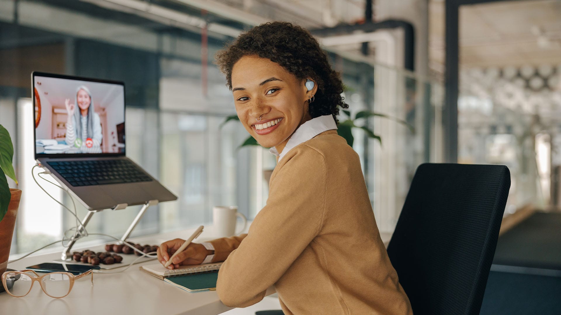 Smiling woman attending a virtual therapy session, writing in a notebook while sitting at a desk.