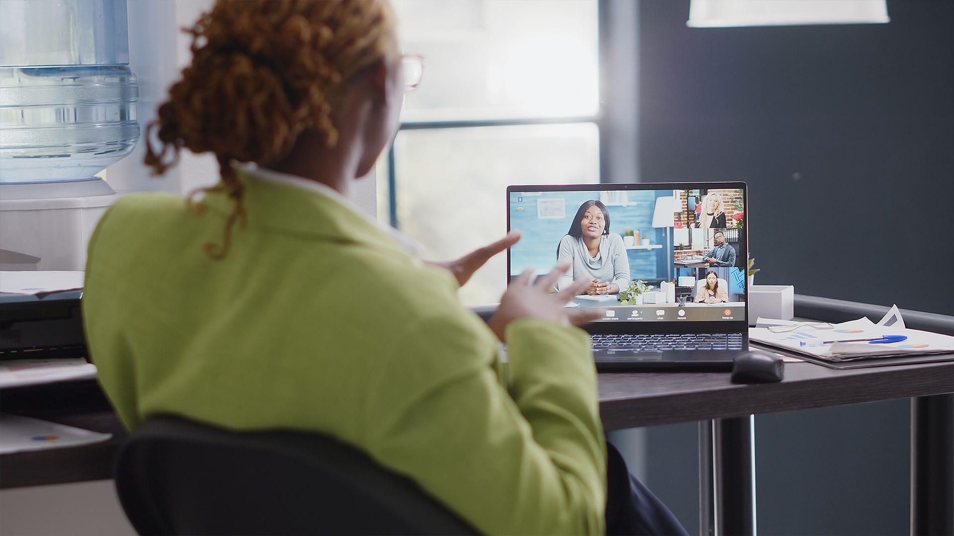 Person sitting at a desk in an office participating in a virtual group meeting, shown on a laptop screen with multiple people engaged in conversation.
