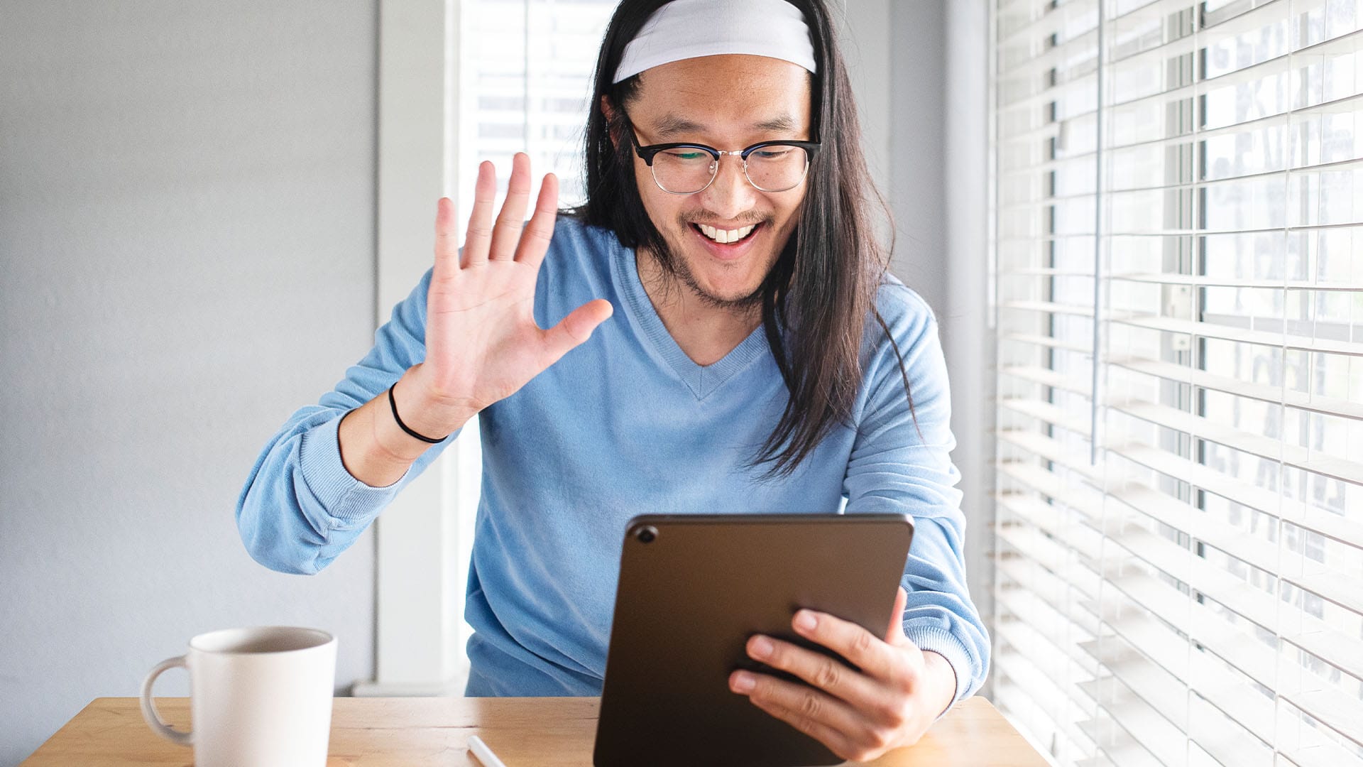 Smiling man in a blue sweater and headband waving at a tablet during a video call.