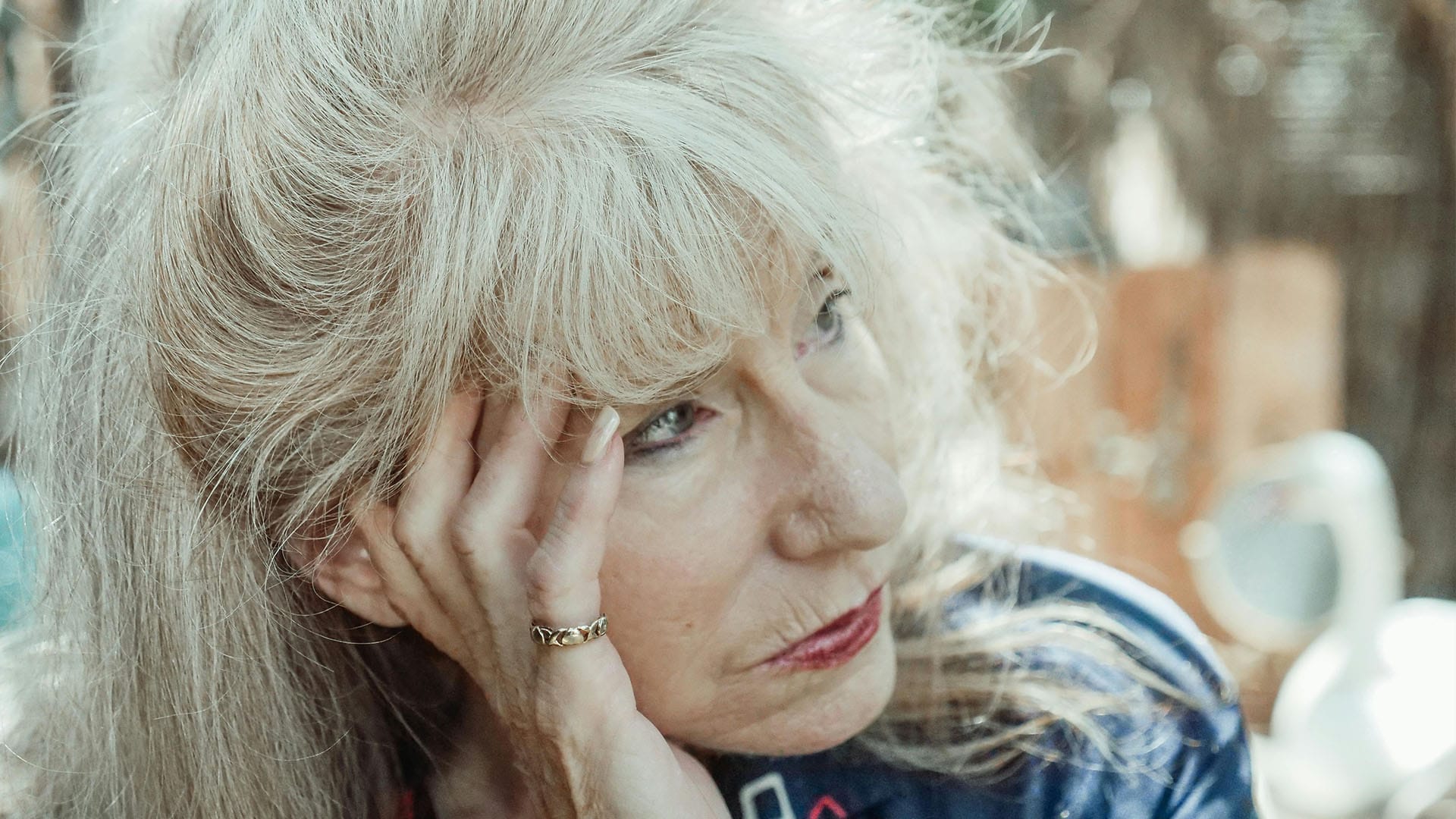 An older woman with long silver hair rests her head on her hand, gazing into the distance with a tired, contemplative expression.