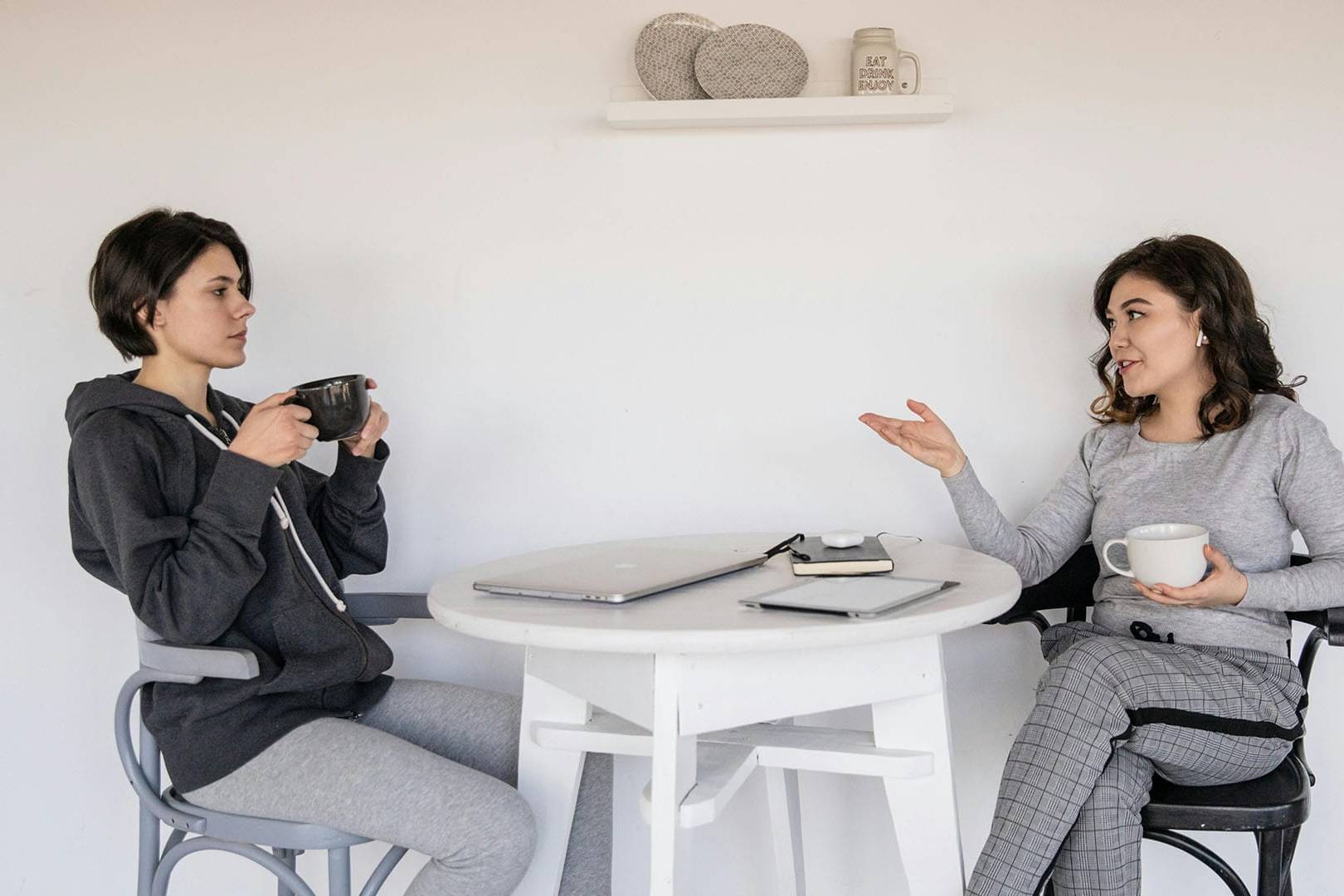 Two women sit across from each other at a small table with mugs in hand, having a supportive and relaxed conversation in a bright, minimalistic setting.