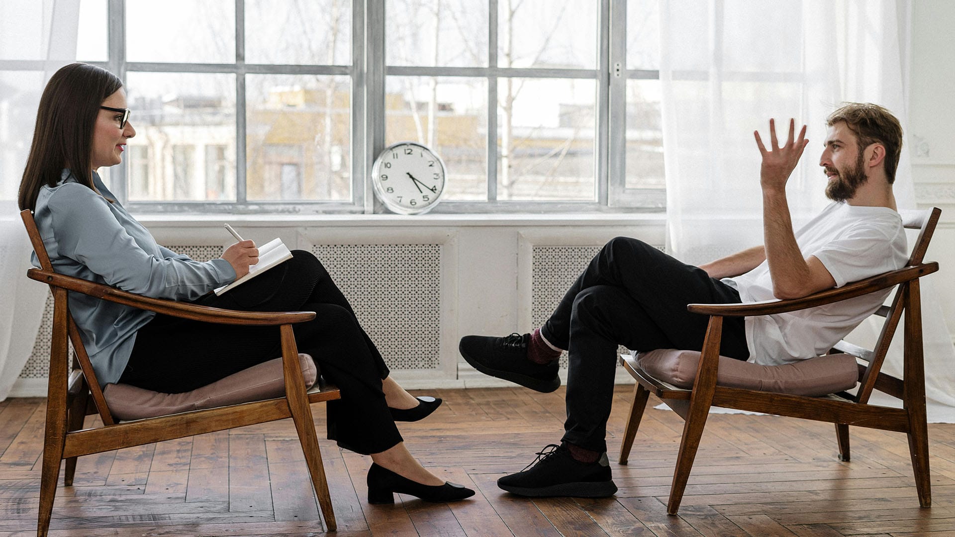 A male client gestures while speaking to a female therapist who is taking notes during a session in a modern, sunlit office with large windows.