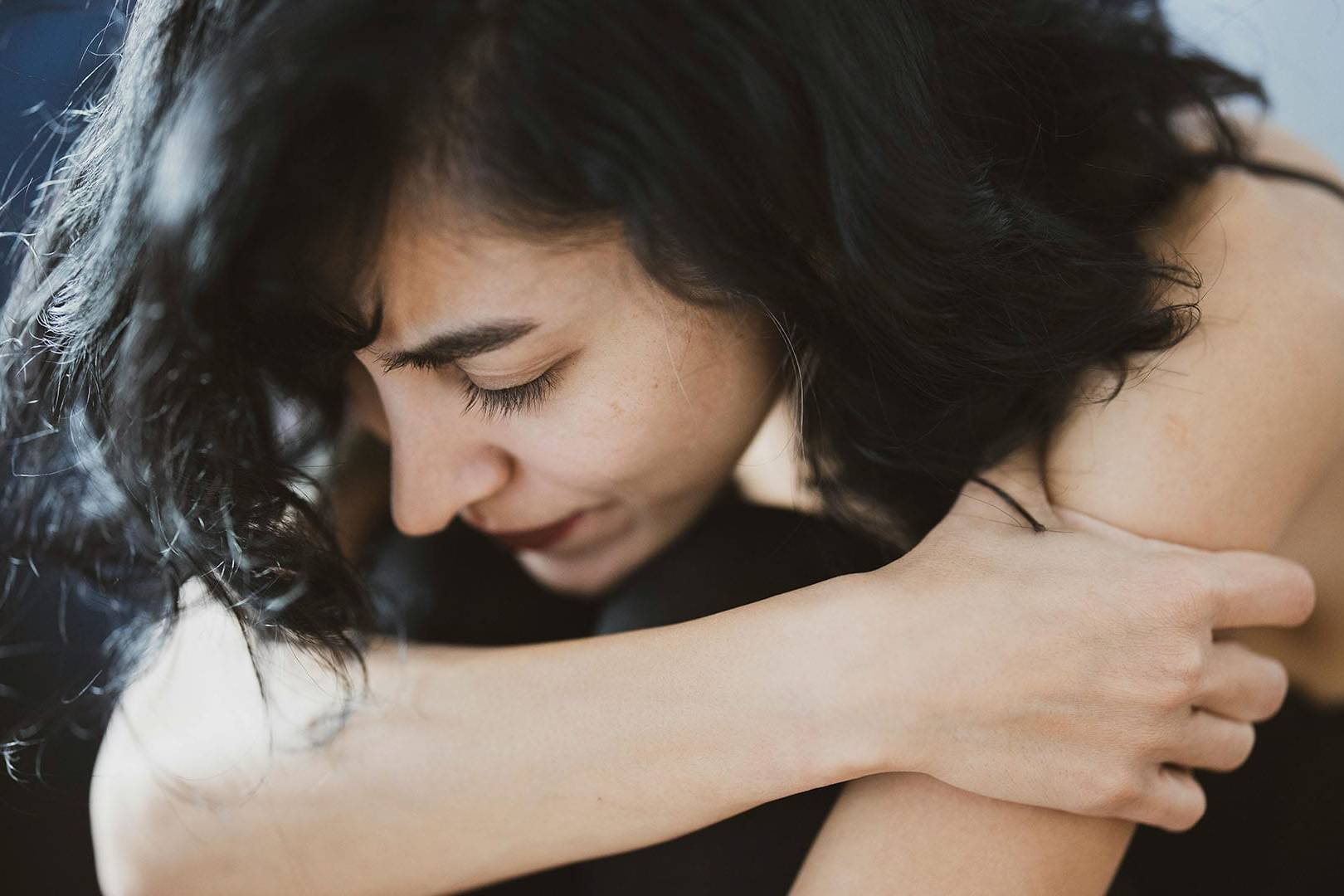 A woman sits curled up with her eyes closed and arms wrapped around her knees, expressing emotional pain, introspection, or sadness.