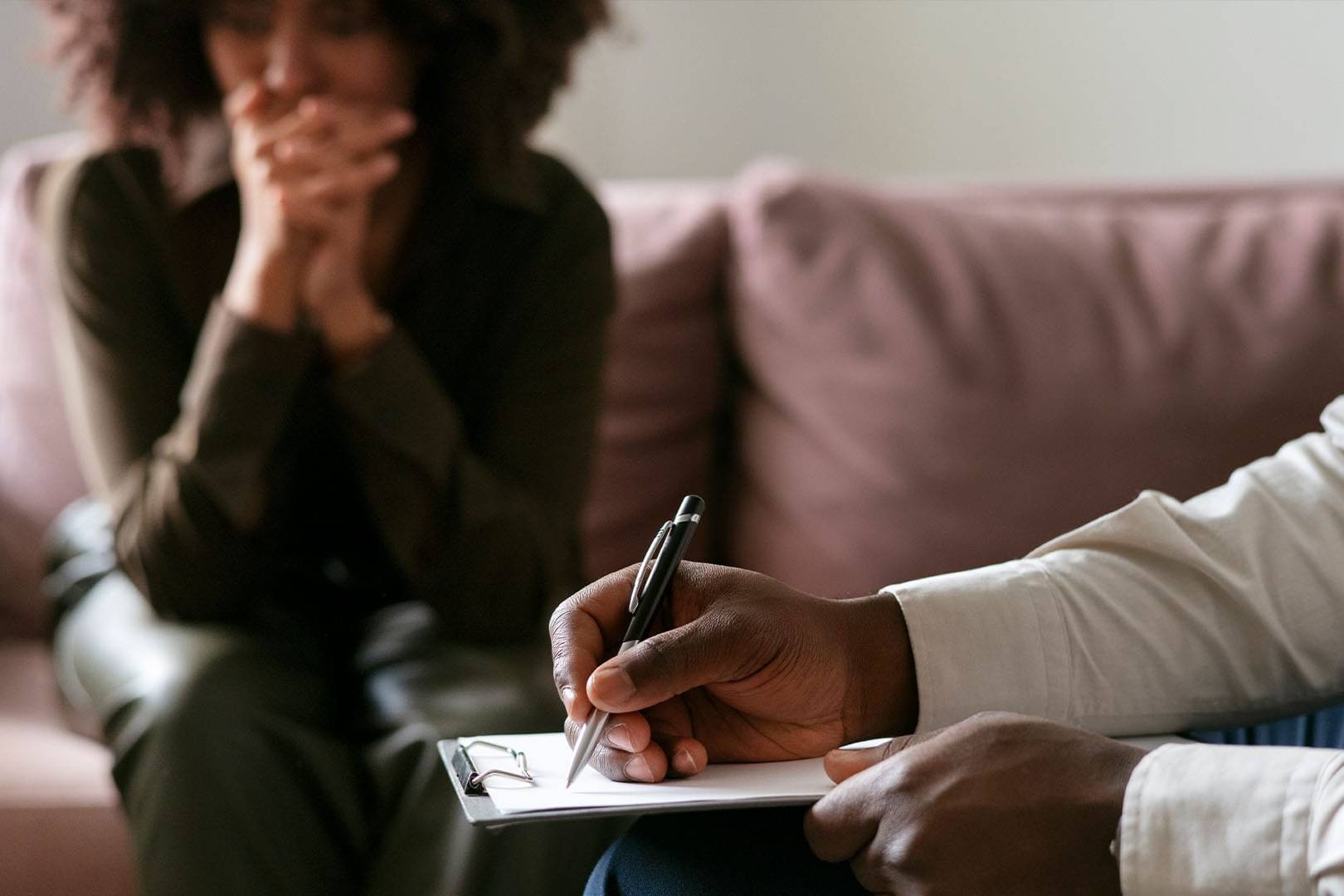 A therapist takes notes while a woman sits on a couch with her hands clasped, appearing anxious or deep in thought during a therapy session.
