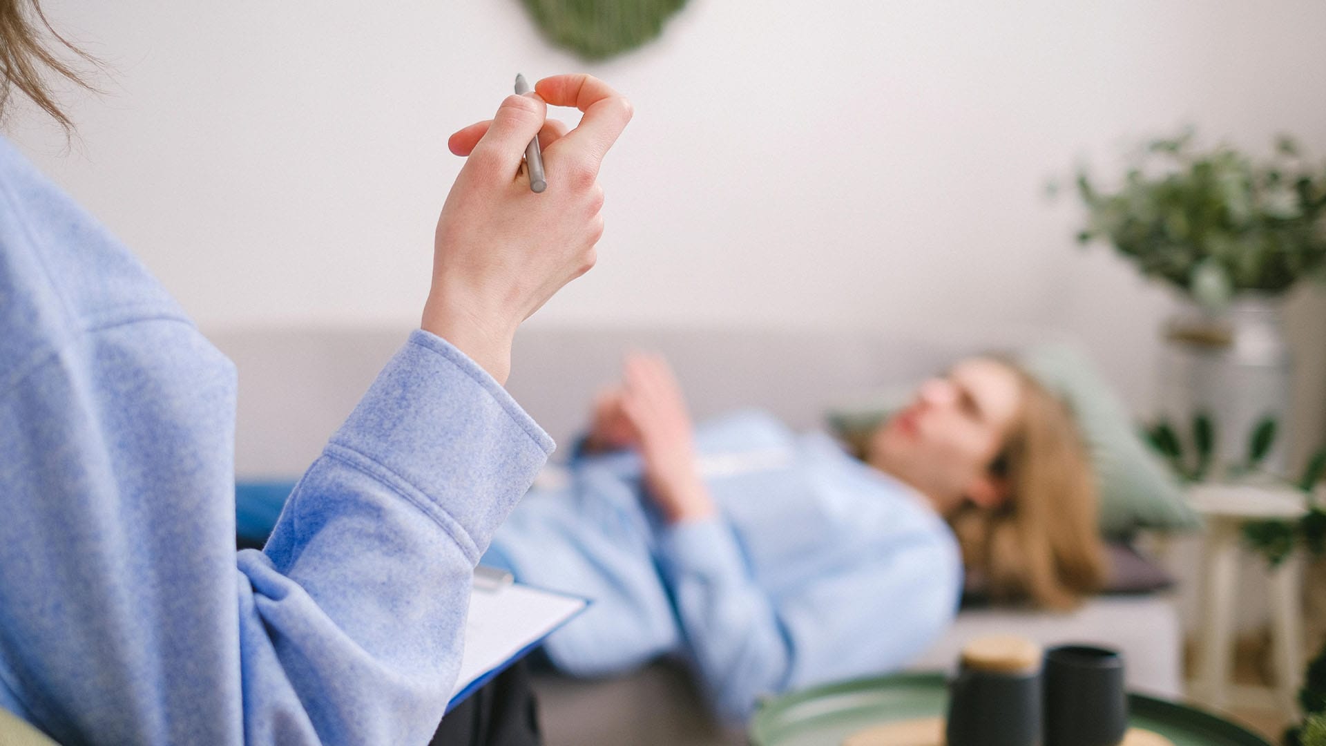 Close-up of therapist’s hand holding a pen while a young adult male lies on a therapy couch during a reflective session in a softly lit room.