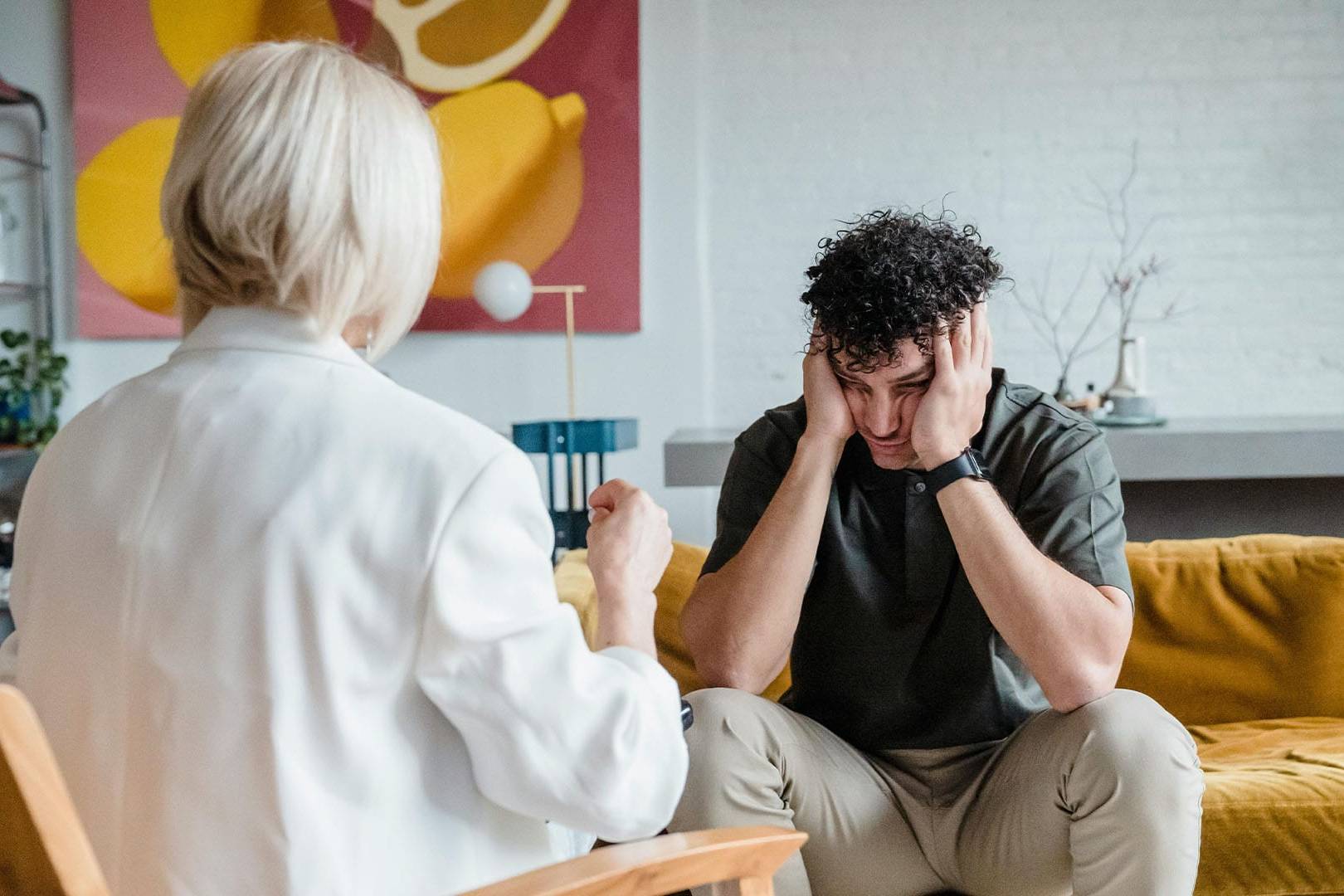 An overwhelmed parent holds his head in his hands while speaking with a therapist in a calm, modern therapy office.