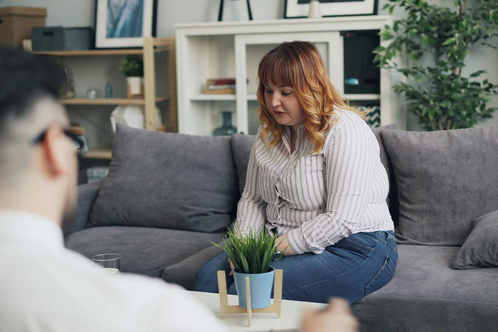 Woman seated on a gray couch during a therapy session, appearing reflective while speaking to an unseen therapist in a calm, professional setting.