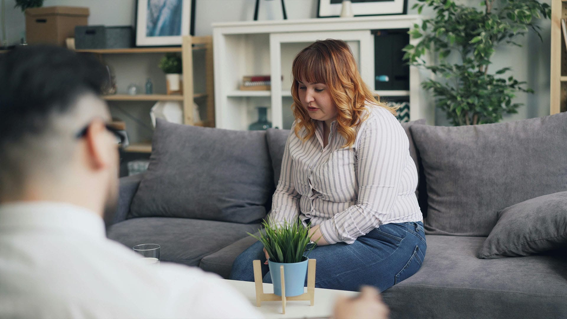 Woman seated on a gray couch during a therapy session, appearing reflective while speaking to an unseen therapist in a calm, professional setting.