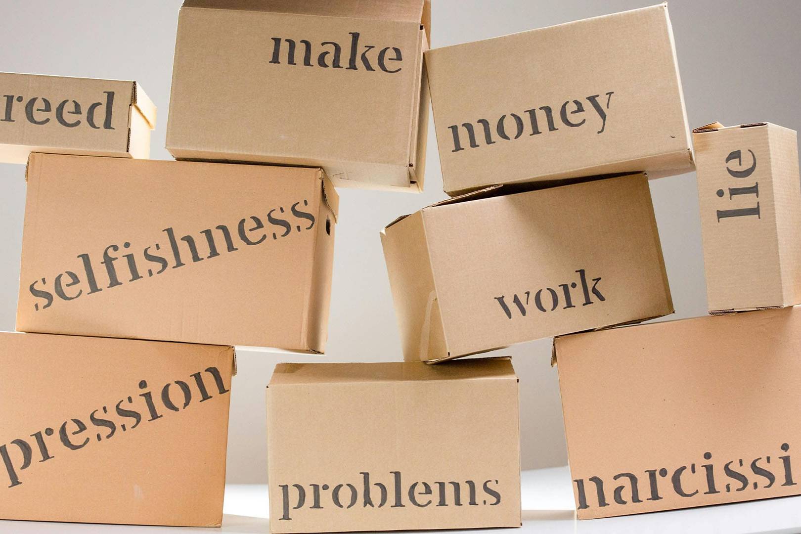 Stacked cardboard boxes labeled with words such as “depression,” “problems,” “greed,” “selfishness,” “lie,” “narcissism,” and “work,” symbolizing emotional and psychological baggage.
