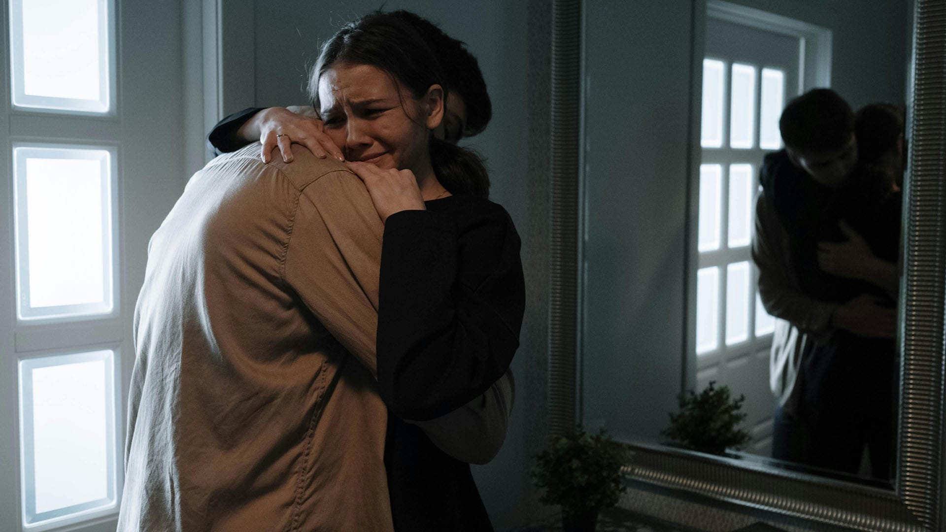 A distressed woman clings to a man in a dimly lit room, visibly crying and overwhelmed, as their reflection appears in a nearby mirror.