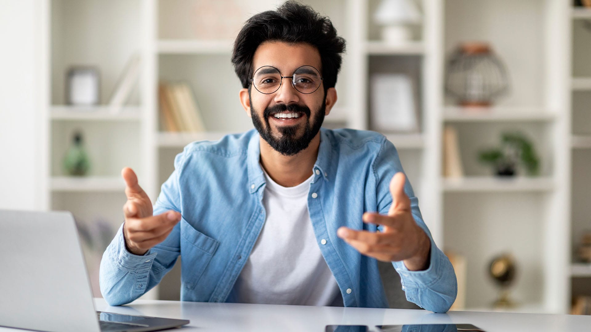 Smiling man in glasses speaking during a video call, seated at a desk in a bright room.