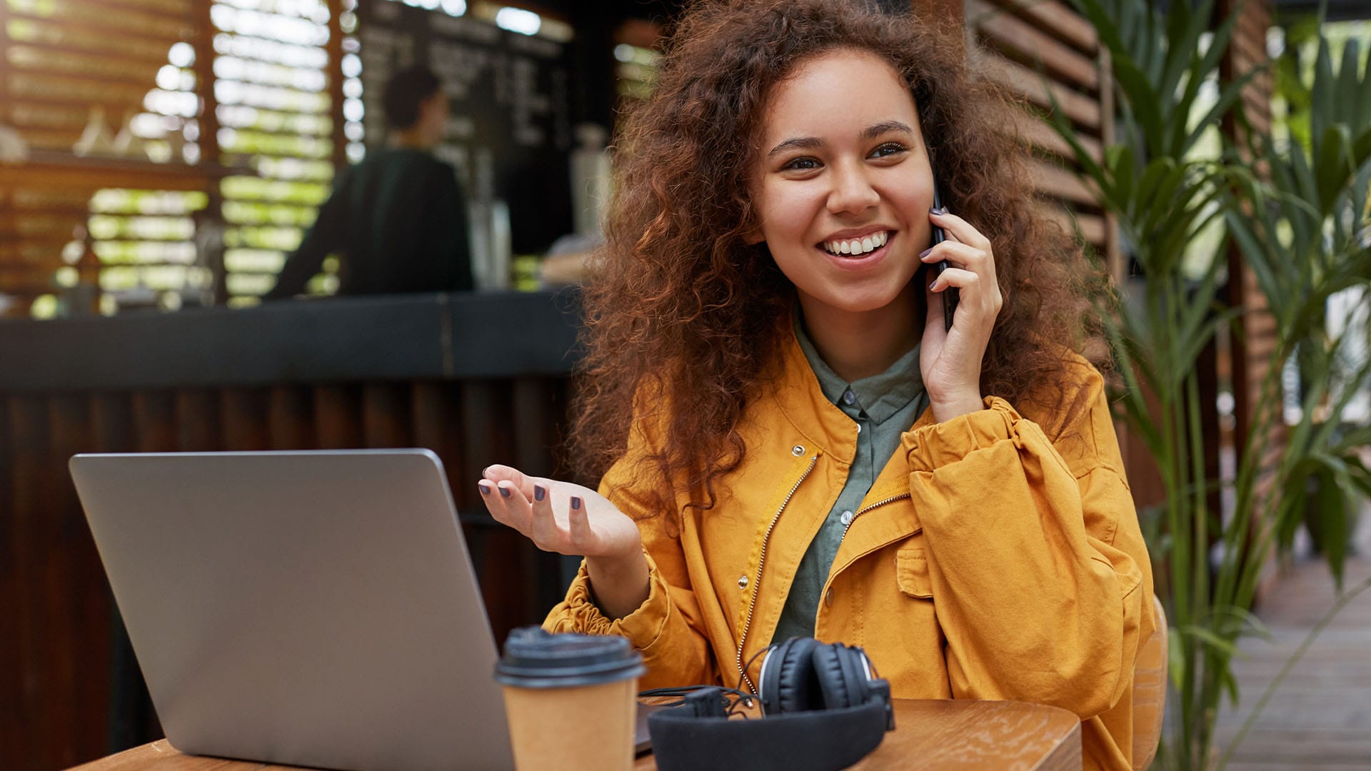 Young woman smiling and talking on the phone while working on a laptop in an outdoor café, symbolizing connection and empowerment.