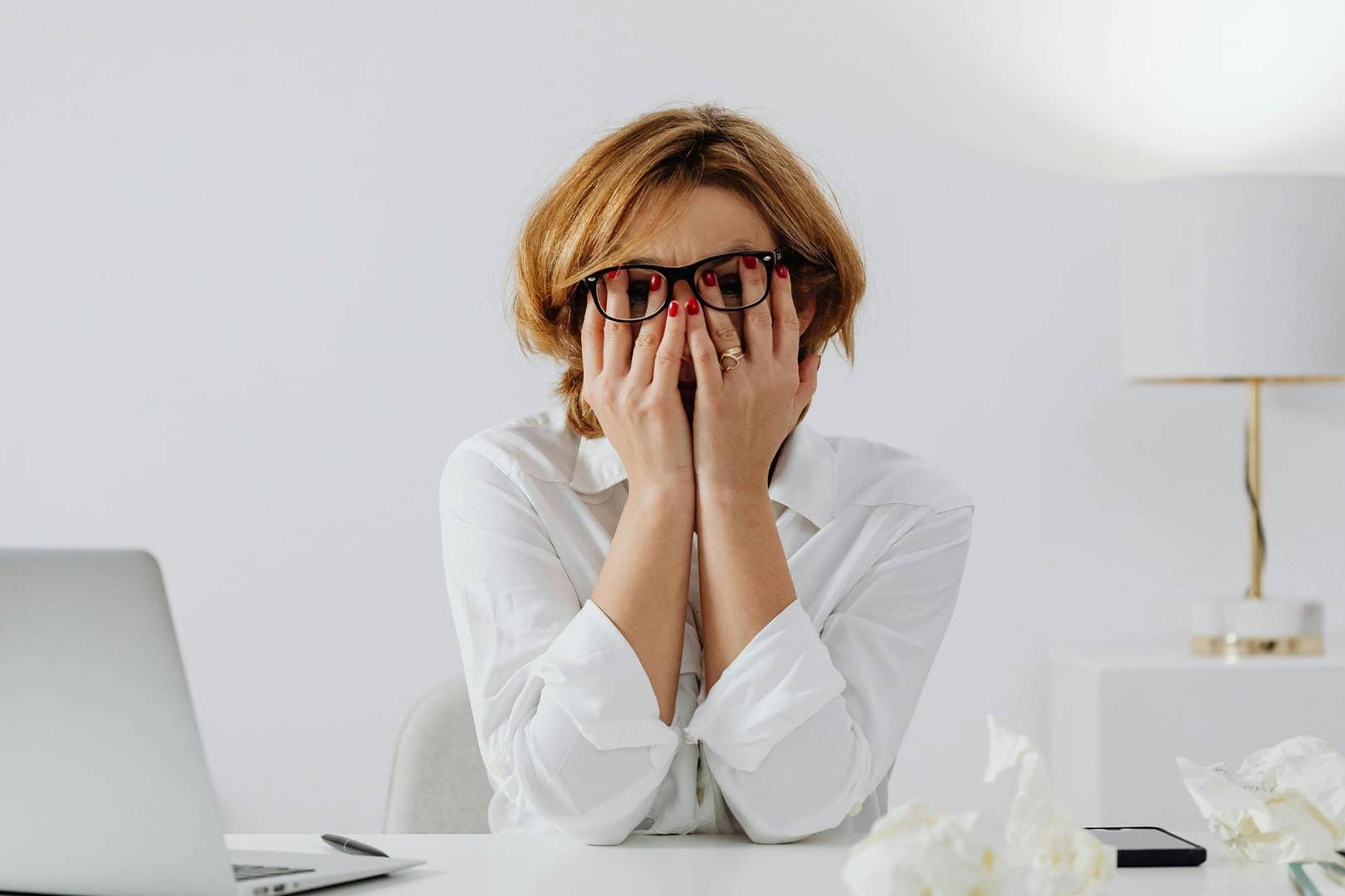 A distressed woman with red nails covers her face with both hands while sitting at a desk with a laptop and crumpled papers, suggesting mental or emotional fatigue.