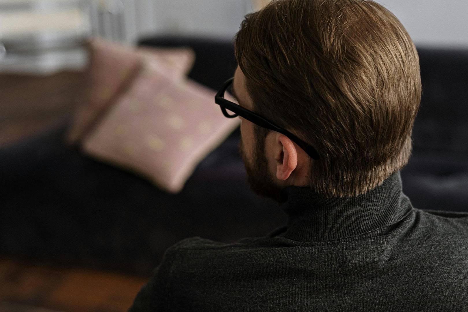 Man in glasses sits on a couch, facing away, in a calm and introspective setting.