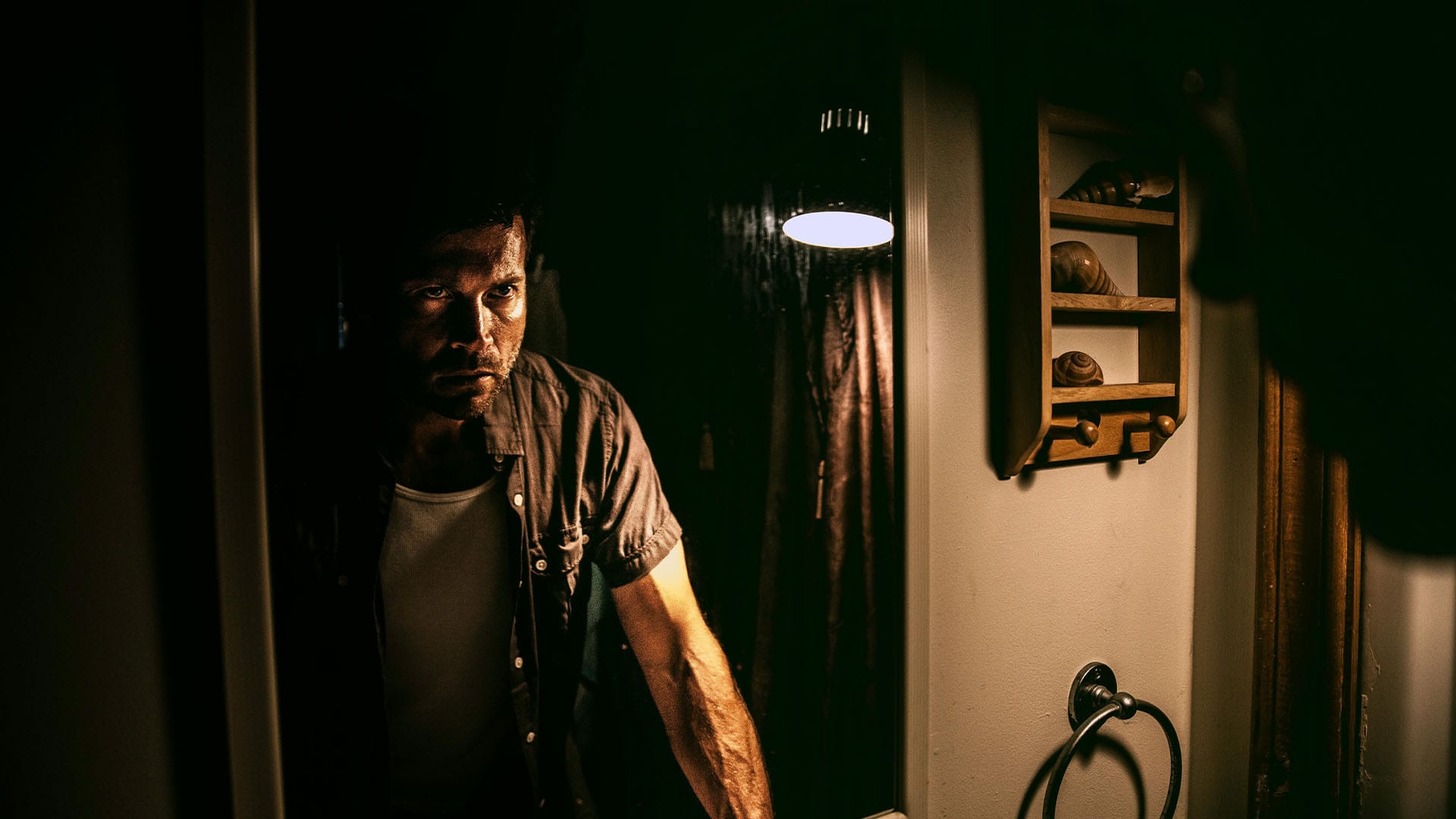 A man stares into a bathroom mirror under dim lighting, gripping the sink with tense posture, reflecting exhaustion, frustration, or burnout.