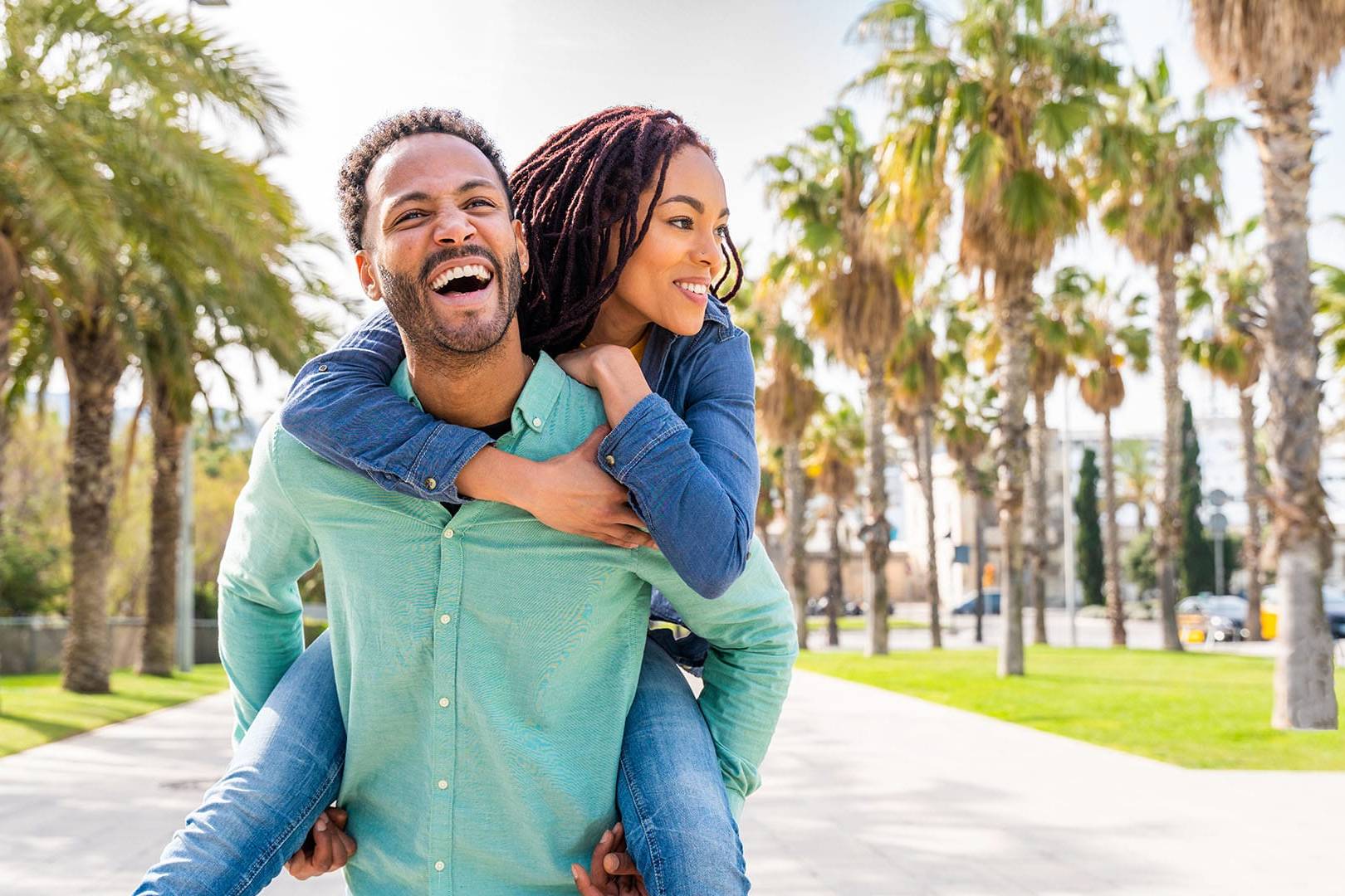 Happy couple enjoying a piggyback ride outdoors, surrounded by palm trees on a sunny day.