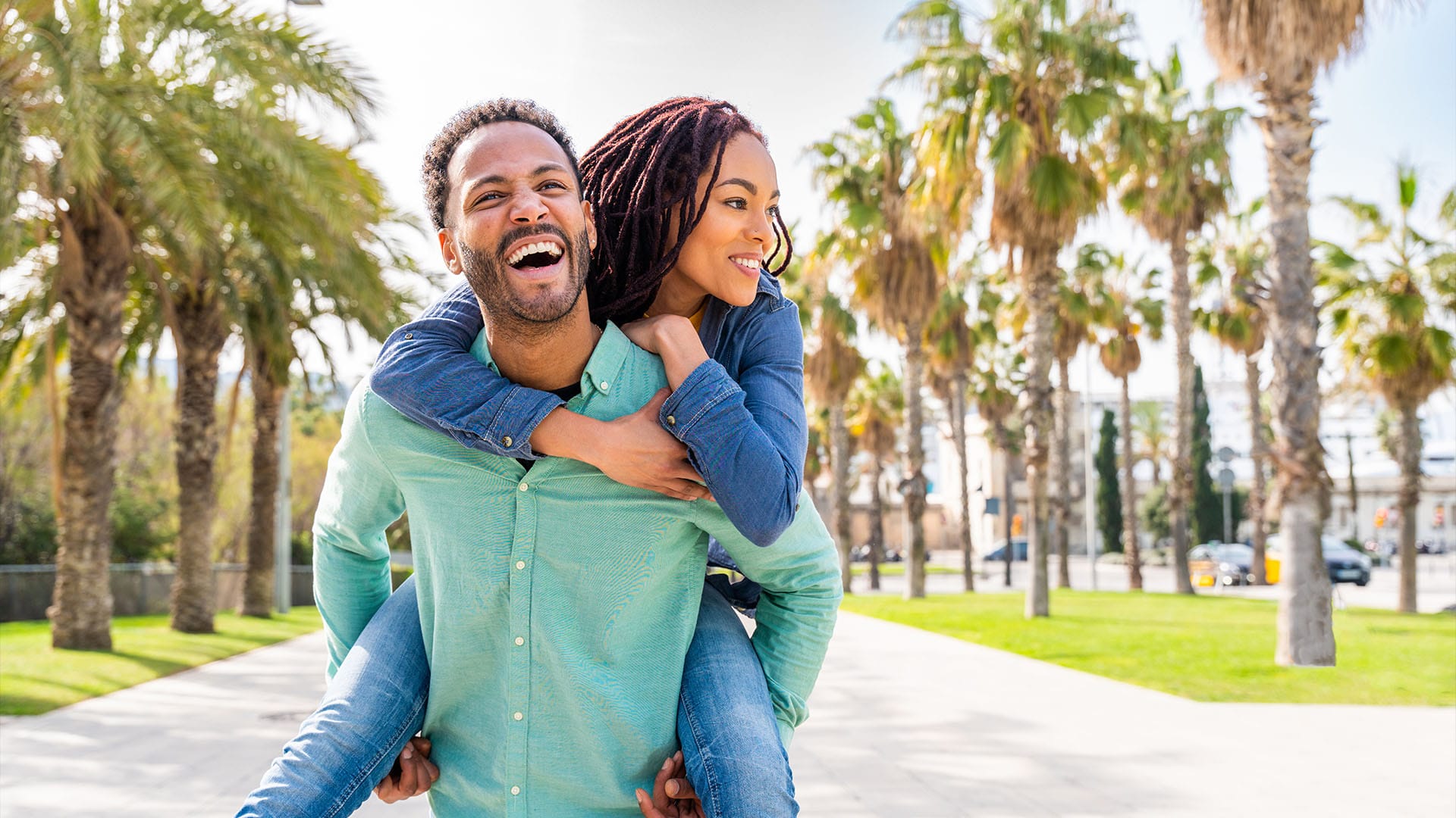 Happy couple enjoying a piggyback ride outdoors, surrounded by palm trees on a sunny day.