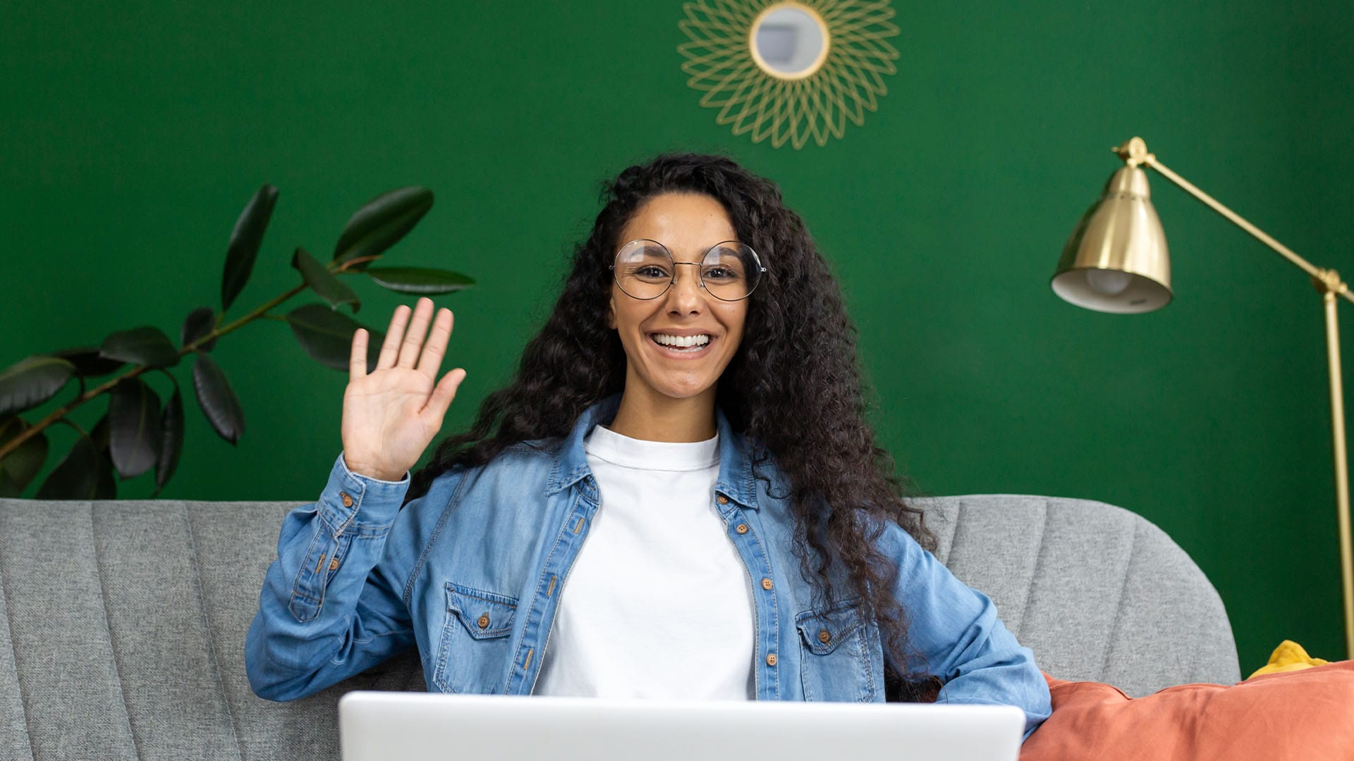 Smiling woman with long curly hair waving at her laptop during a virtual session, seated on a couch with a green background.